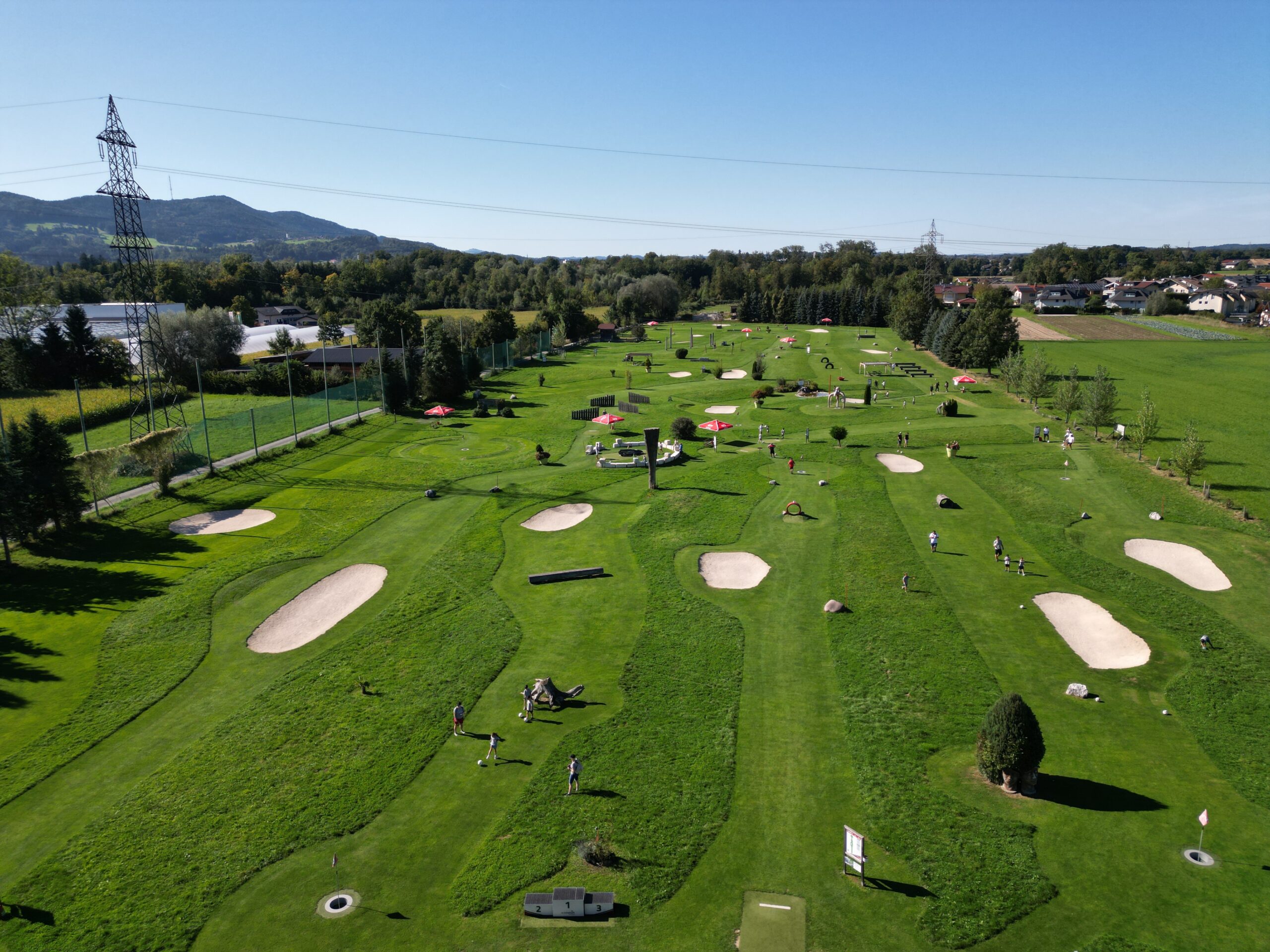Luftaufnahme eines gr&uuml;nen Golfplatzes mit Sandbunkern, B&auml;umen und Menschen, die unter strahlend blauem Himmel spielen.