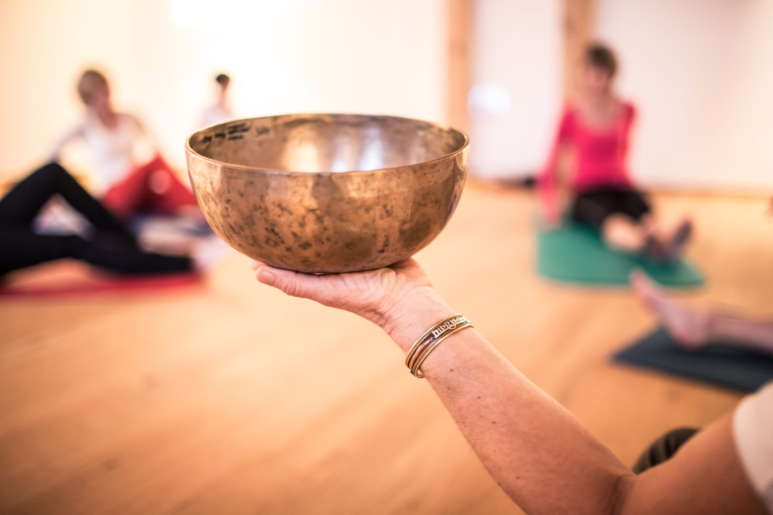 Im Vordergrund ist eine Hand zu sehen, die eine Klangschale h&auml;lt; im unscharfen Hintergrund sitzen Menschen auf Yogamatten.
