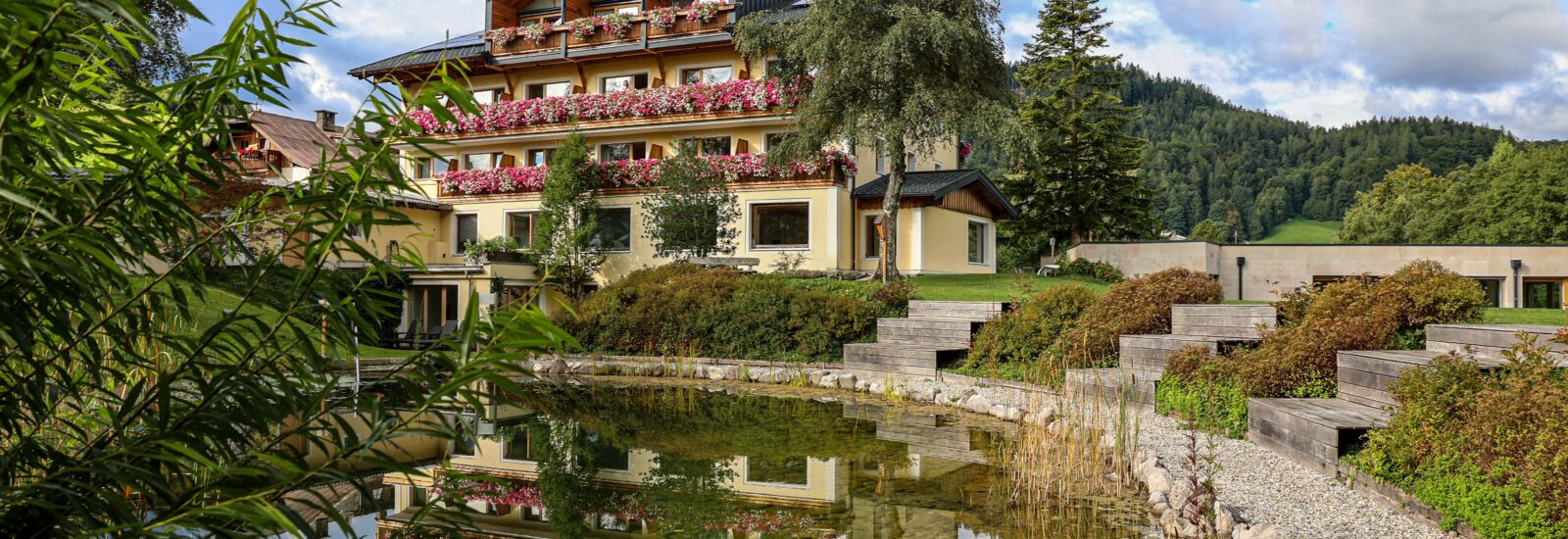 Ein Hotel am See mit Balkonen, Blumenkästen und einem Blick auf die Berge, die sich im Teich spiegeln.