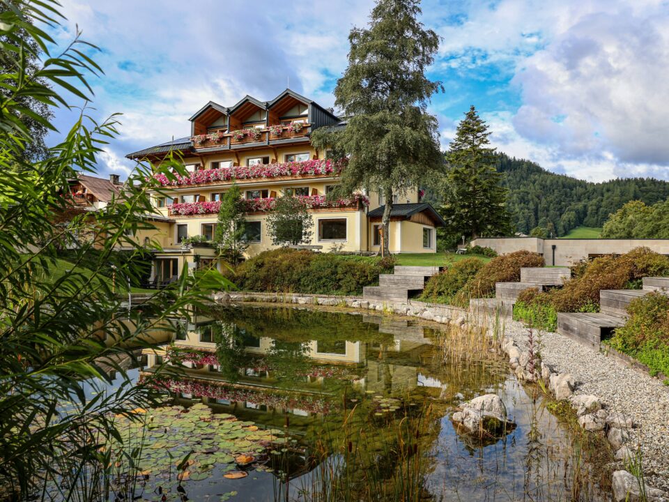Ein Hotel am See mit Balkonen, Blumenkästen und einem Blick auf die Berge, die sich im Teich spiegeln.