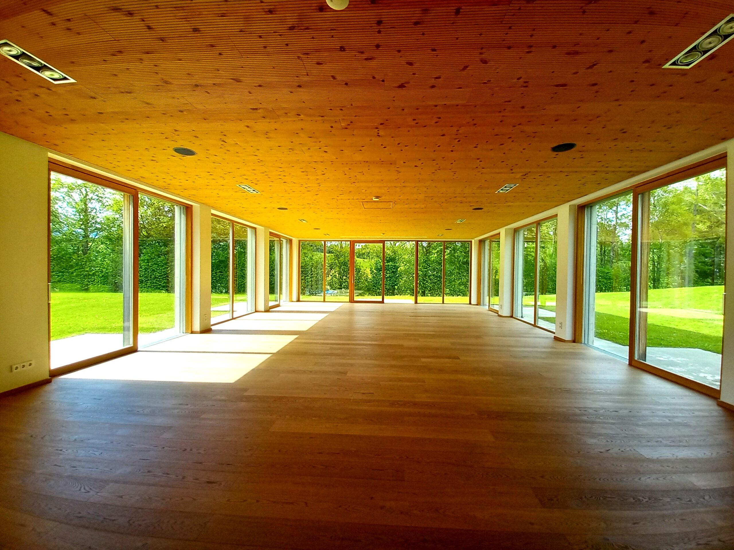 Ein heller, leerer Raum mit Holzboden und Holzdecke, umgeben von gro&szlig;en Glasfenstern mit Blick auf gr&uuml;ne Rasenfl&auml;chen.