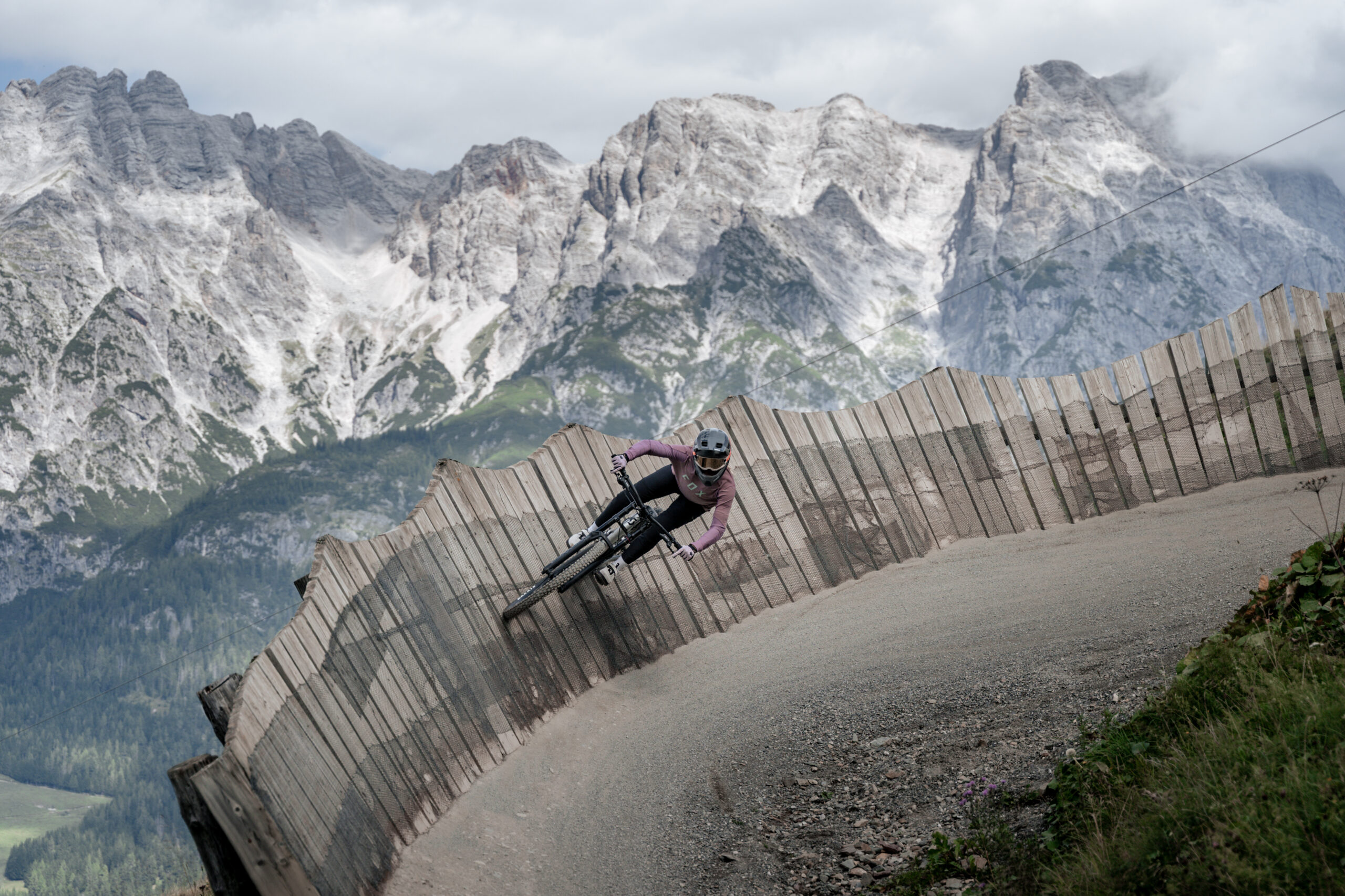 Ein Mountainbiker fährt auf einer kurvigen Holzpiste mit felsigen Bergen im Hintergrund.
