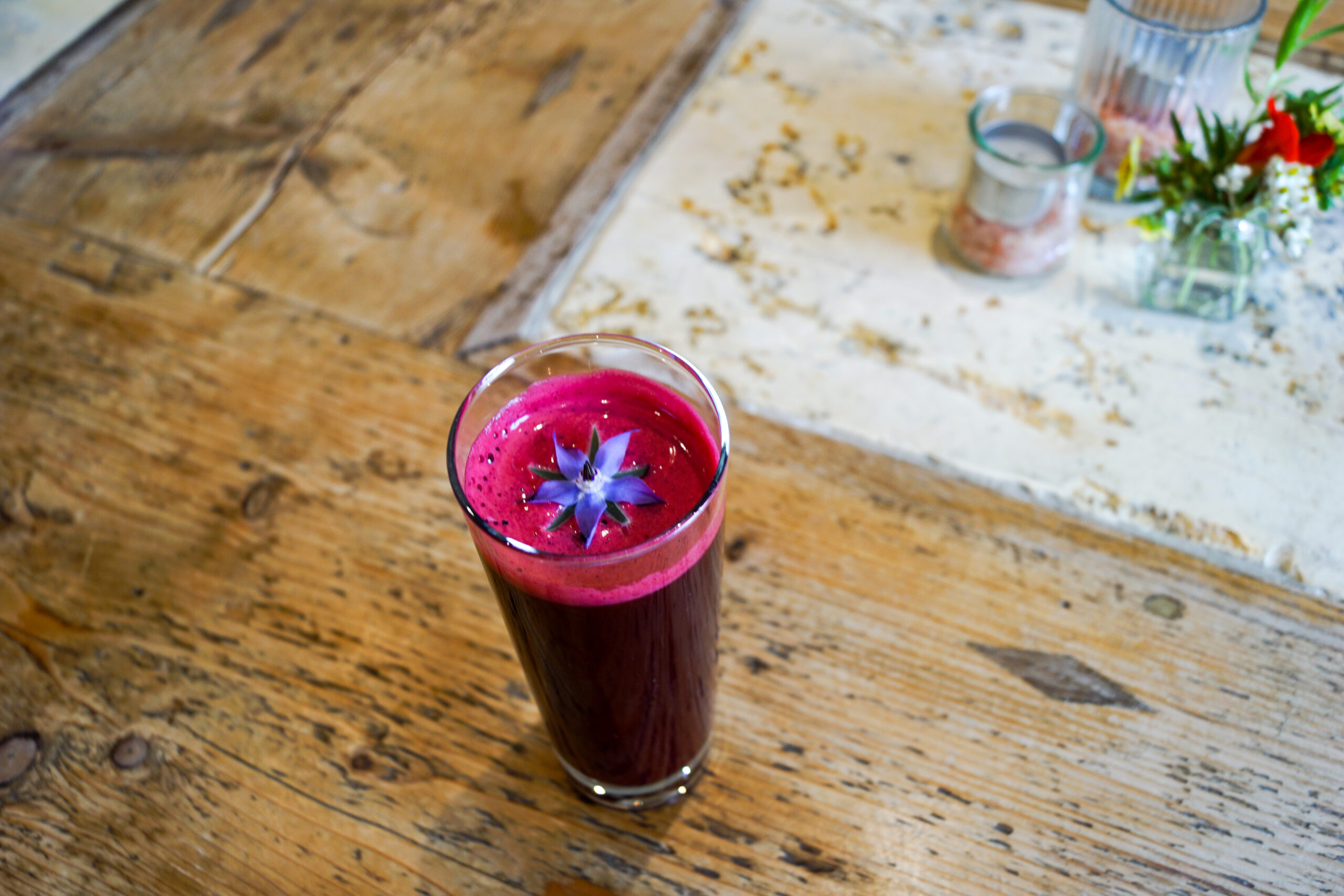Ein Glas dunkelvioletter Saft mit einer violetten essbaren Bl&uuml;te darauf, serviert auf einem rustikalen Holztisch.