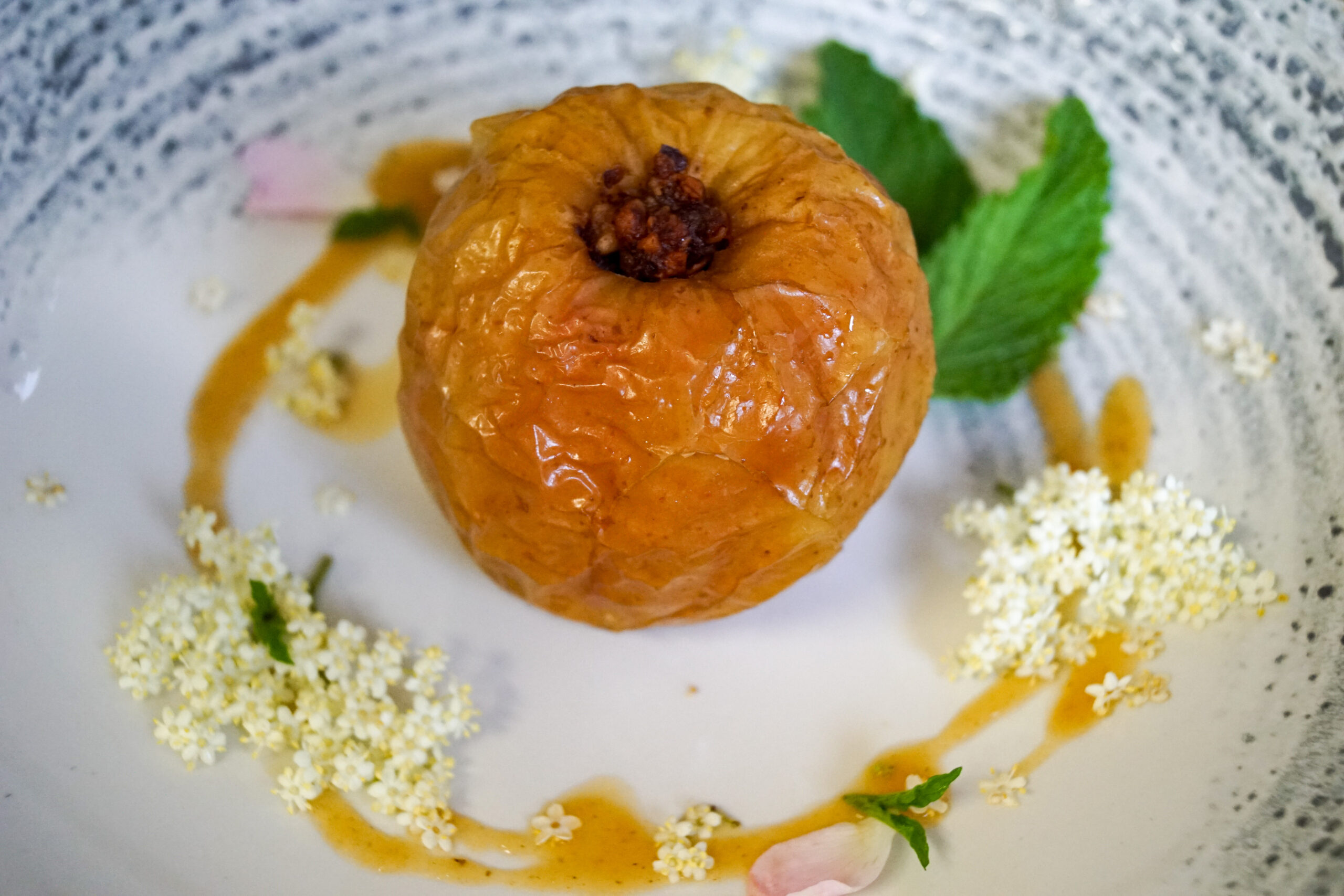 Gebackener Apfel mit So&szlig;e, garniert mit Bl&uuml;ten und Minzbl&auml;ttern auf einem wei&szlig;en Teller.