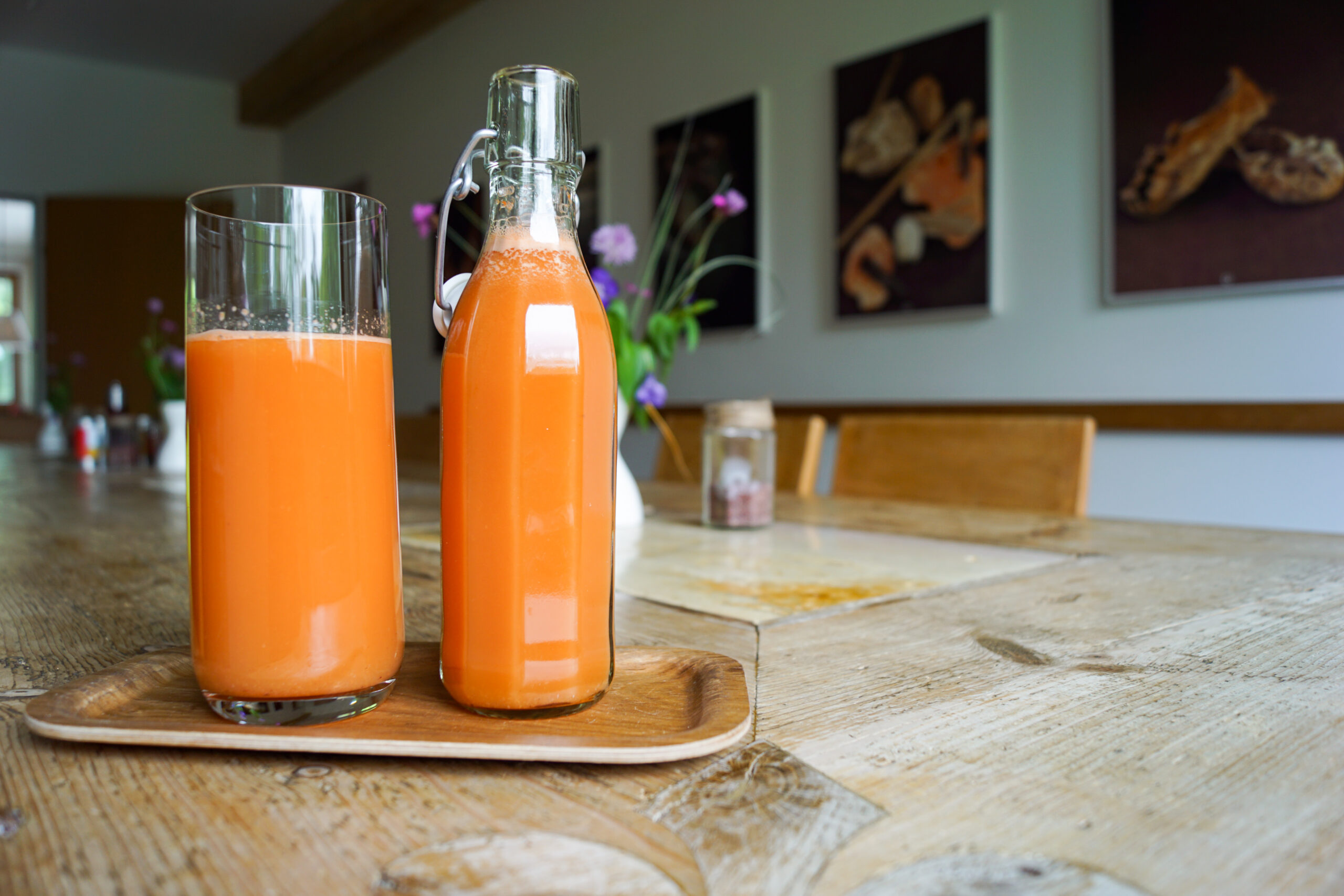 Ein Glas und eine Flasche Orangensaft auf einem Holztablett auf einem rustikalen Esstisch.
