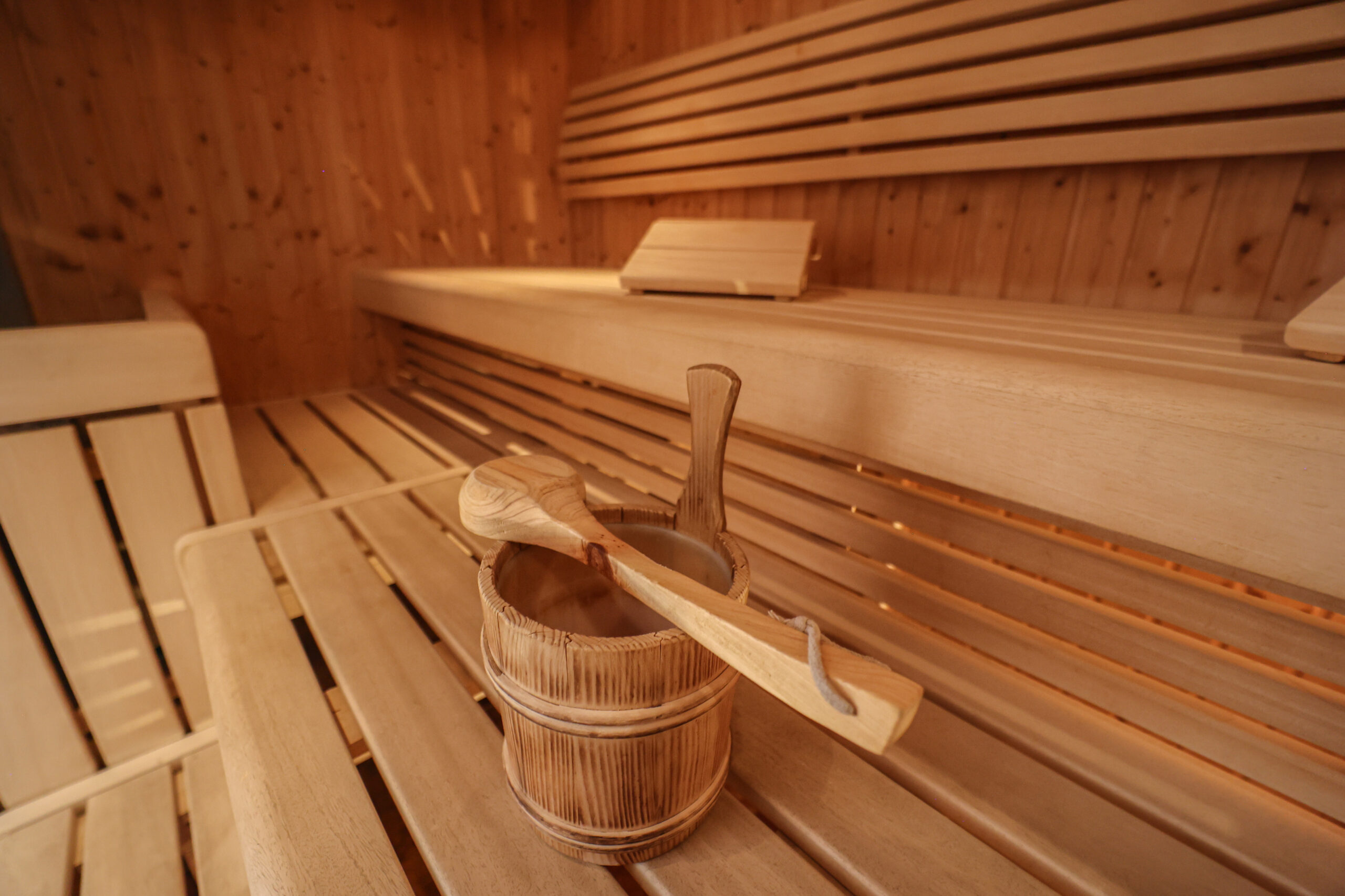 Holzb&auml;nke und Holzw&auml;nde im Inneren einer Sauna, im Vordergrund ein Holzeimer und eine Sch&ouml;pfkelle auf einer Bank.