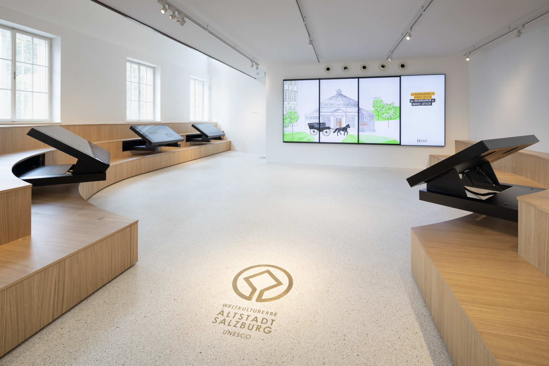 Moderner Ausstellungsraum mit interaktiven Bildschirmen, Holzbänken und einer großen digitalen Anzeige über das UNESCO-Weltkulturerbe in Salzburg.