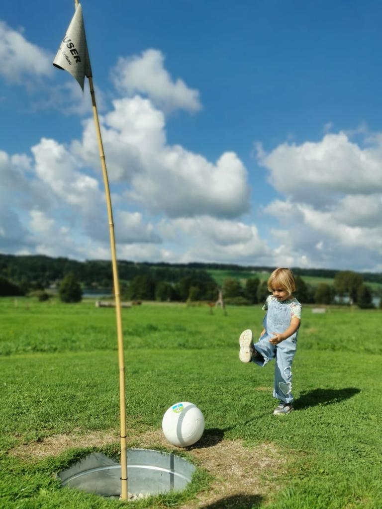 Ein kleines Kind kickt einen gro&szlig;en Ball auf einem Rasenfeld unter blauem Himmel in Richtung eines Lochs unter einer Flagge.