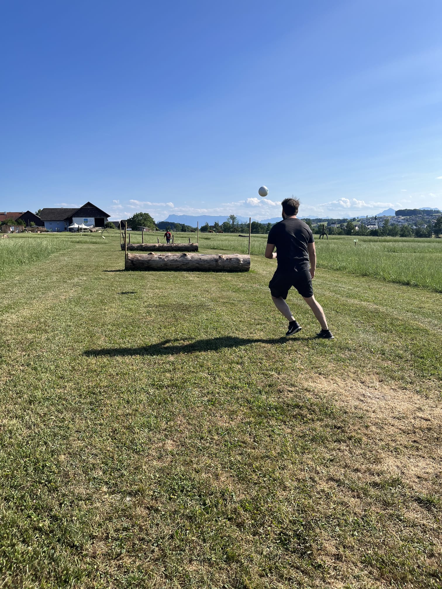 Eine Person spielt im Freien unter strahlend blauem Himmel auf einer Wiese in der N&auml;he von Baumstammhindernissen mit einem Ball.