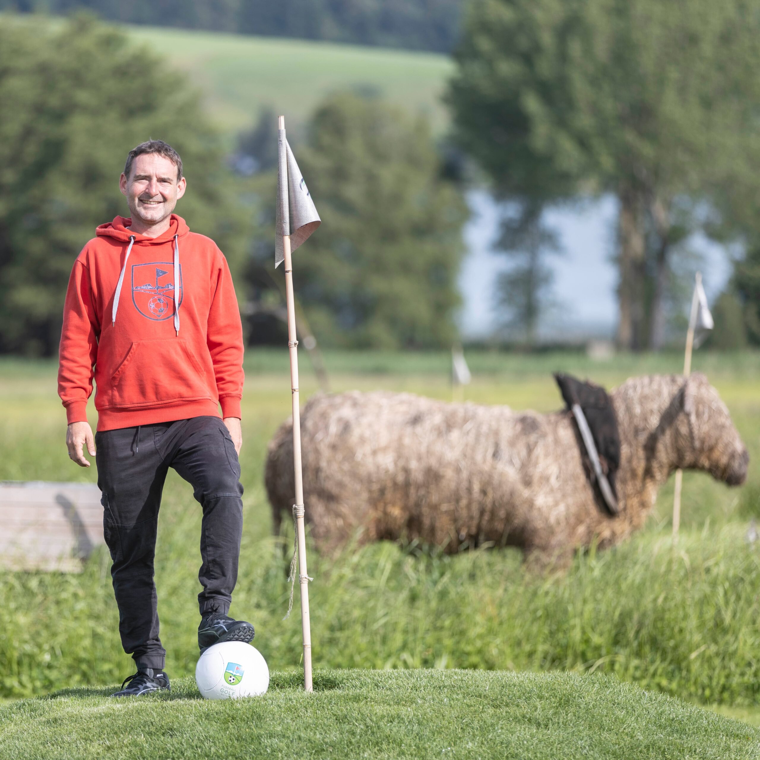 Ein Mann in einem roten Kapuzenpulli steht auf dem Rasen, einen Fu&szlig; auf einem Fu&szlig;ball, neben einer Flagge und einer Heuskulptur.