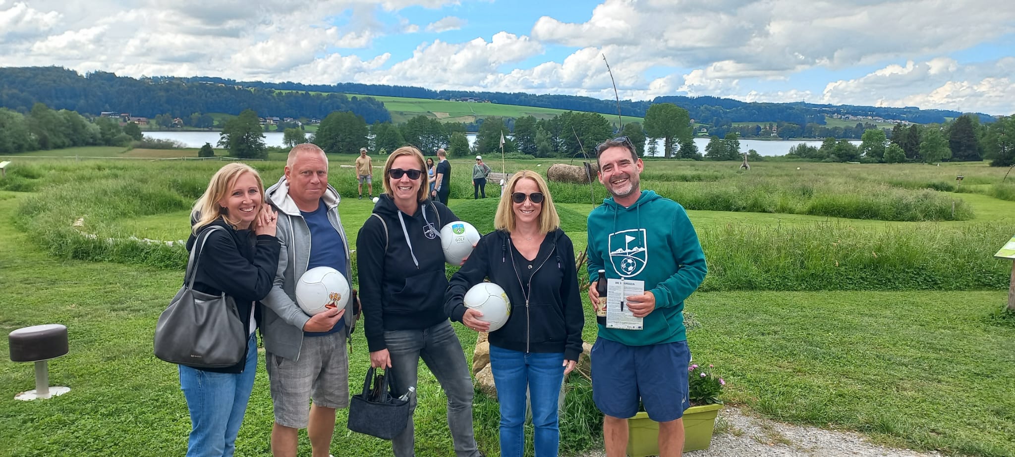 F&uuml;nf Menschen stehen l&auml;chelnd drau&szlig;en auf einer Wiese, halten Fu&szlig;b&auml;lle in den H&auml;nden, im Hintergrund sind ein See und H&uuml;gel zu sehen.