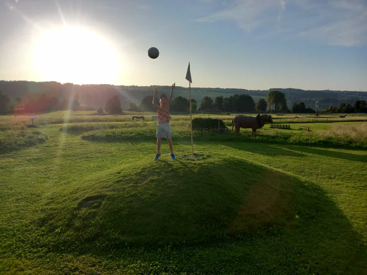 Ein Kind wirft einen Ball in der N&auml;he einer Fahne auf einem grasbewachsenen H&uuml;gel auf einem sonnigen Feld auf dem Land, im Hintergrund ist eine Kuh zu sehen.