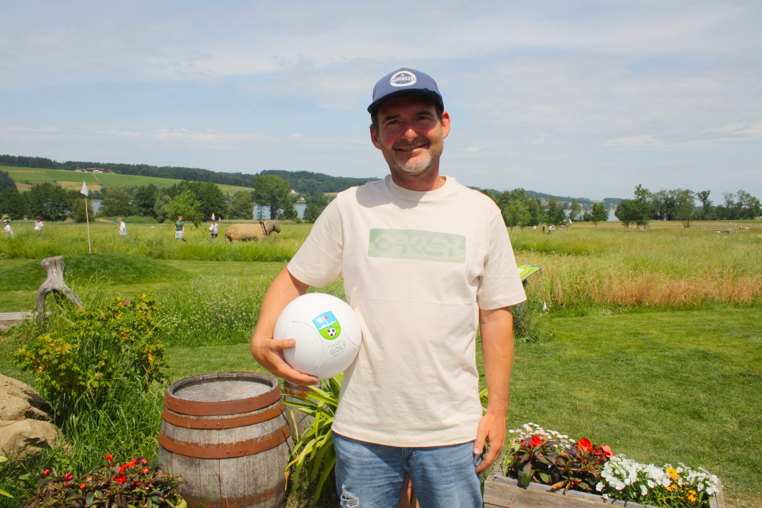 Ein Mann mit einer M&uuml;tze steht im Freien und h&auml;lt einen wei&szlig;en Fu&szlig;ball in der Hand; im Hintergrund sind Gras, Blumen und Felder zu sehen.