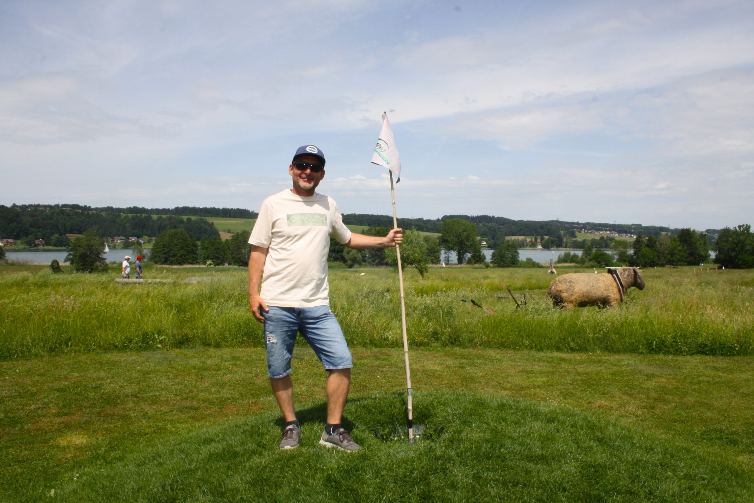 Ein Mann in Freizeitkleidung steht neben einer Golffahne auf einer Wiese, im Hintergrund sind ein See und B&auml;ume zu sehen.