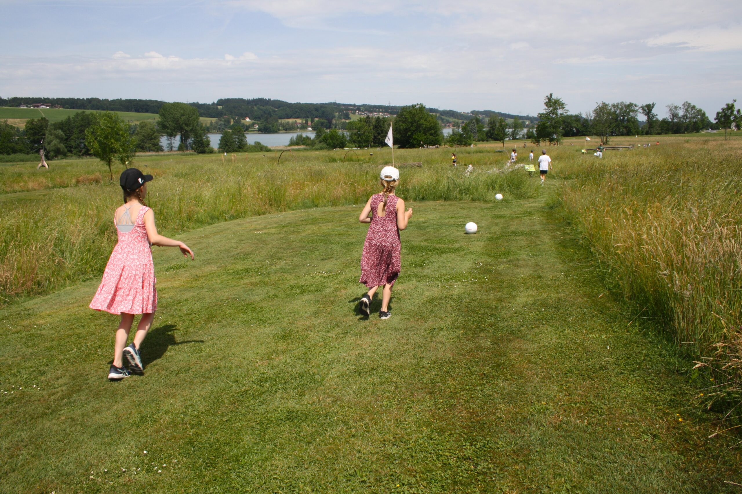 Kinder in Sommerkleidung spielen unter einem teilweise bew&ouml;lkten Himmel auf einer Wiese mit gro&szlig;en wei&szlig;en B&auml;llen.