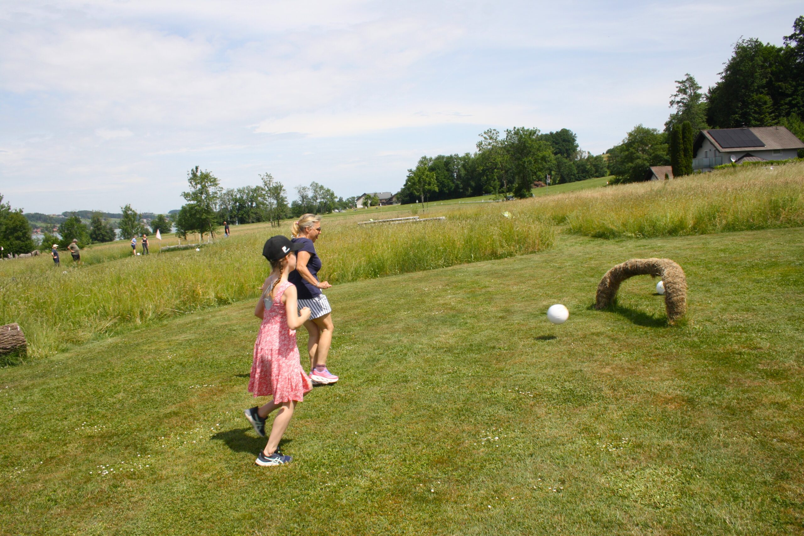 Zwei M&auml;dchen spielen drau&szlig;en Fu&szlig;ball und versuchen, den Ball in einen kleinen Bogen aus Heu auf einer Wiese zu schie&szlig;en.