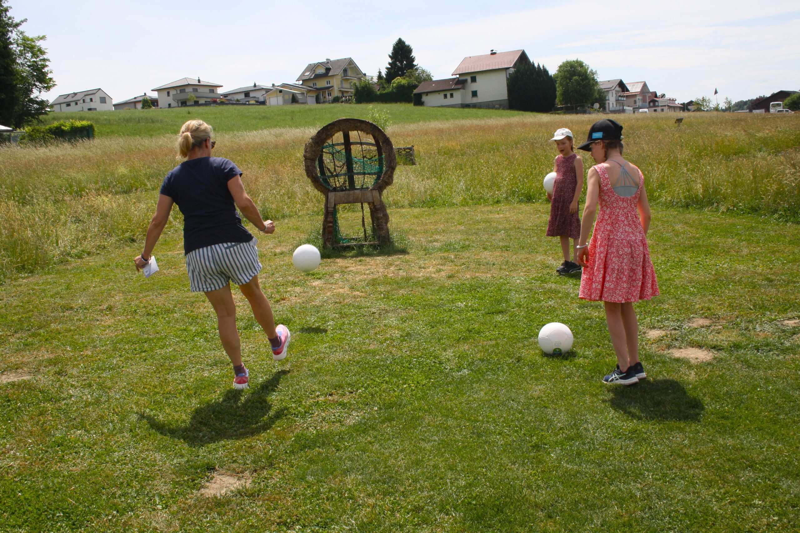 An einem sonnigen Tag spielen drei M&auml;dchen auf einer Wiese in der N&auml;he einer Strohskulptur Fu&szlig;ball.