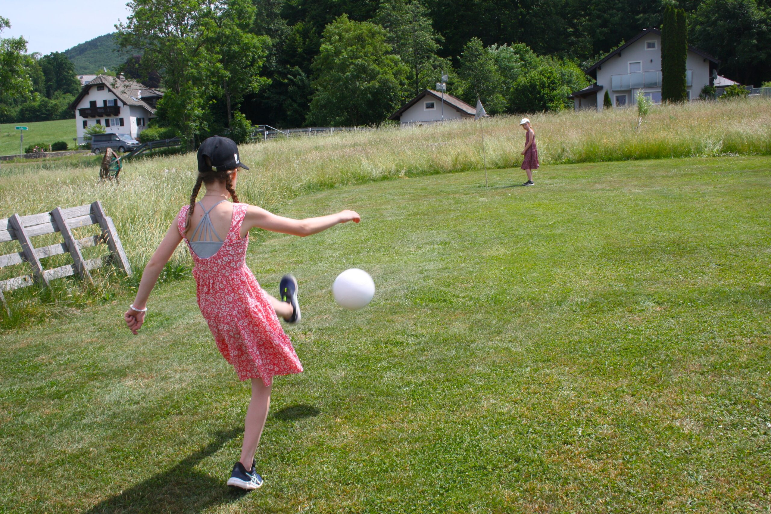Ein M&auml;dchen in einem roten Kleid kickt einen wei&szlig;en Ball in Richtung eines anderen Kindes auf einer Rasenfl&auml;che, im Hintergrund sind H&auml;user zu sehen.