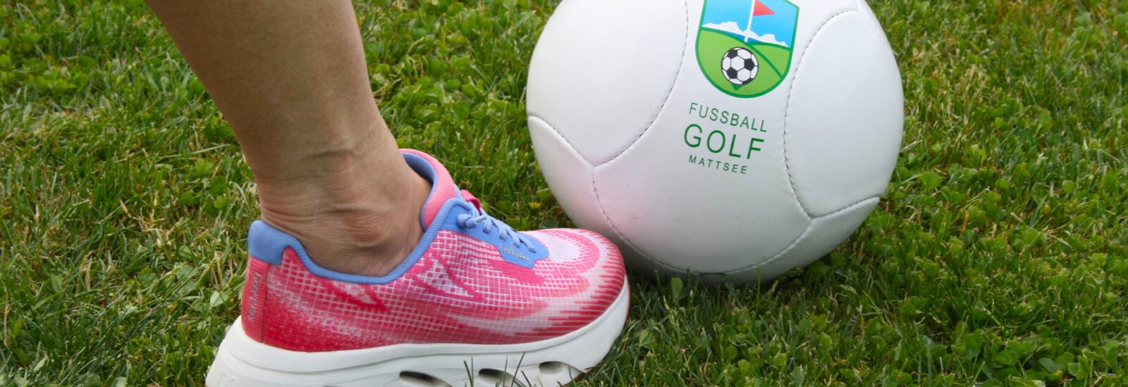 Eine Person in rosa Turnschuhen steht neben einem weißen Fußball auf grünem Rasen.