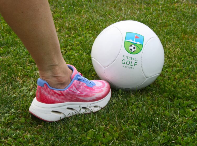 Eine Person in rosa Turnschuhen steht neben einem weißen Fußball auf grünem Rasen.