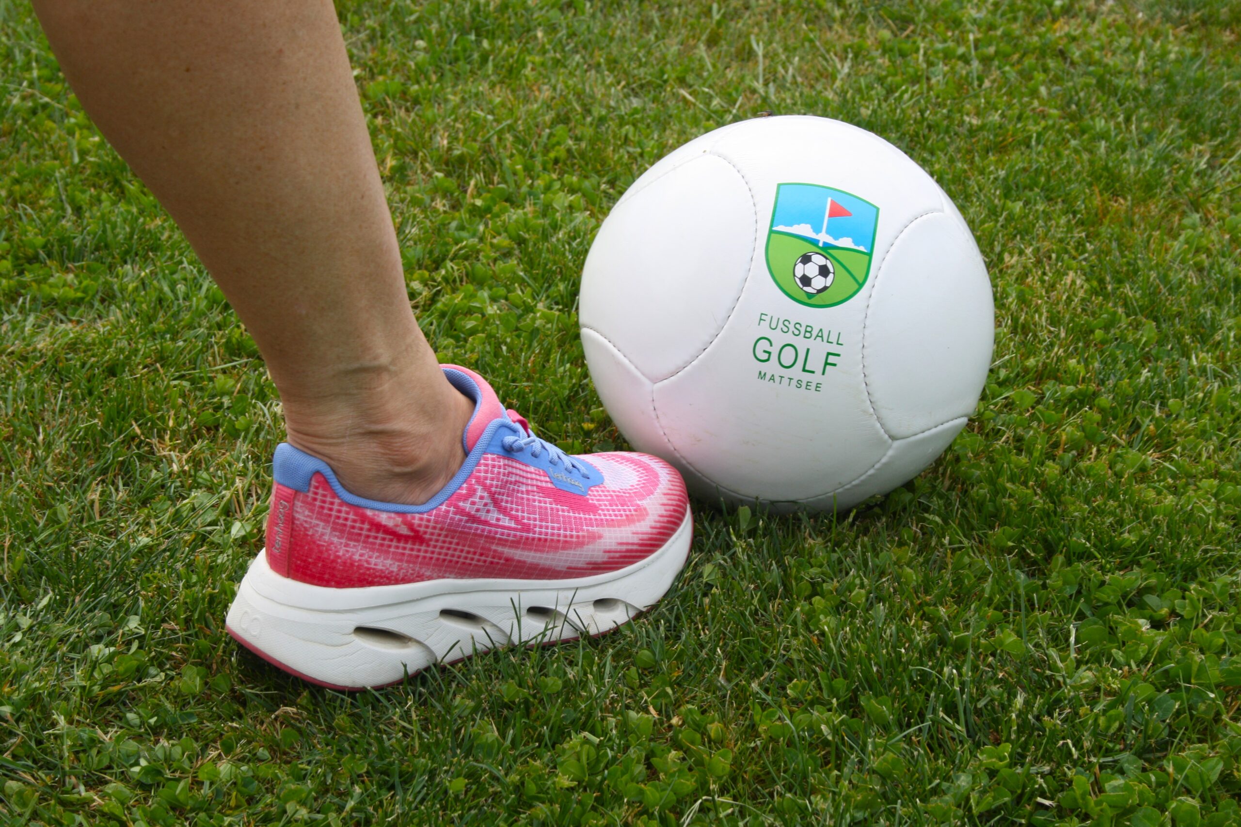 Eine Person in rosa Turnschuhen steht neben einem wei&szlig;en Fu&szlig;ball auf gr&uuml;nem Rasen.