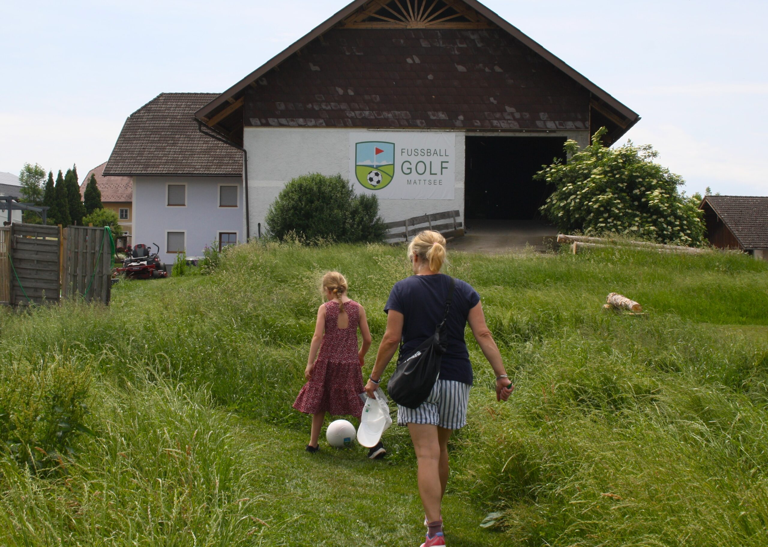 Eine Frau und ein M&auml;dchen gehen auf einem grasbewachsenen Weg auf ein Geb&auml;ude zu, auf dem &bdquo;Fussball Golf Mattsee&ldquo; steht.