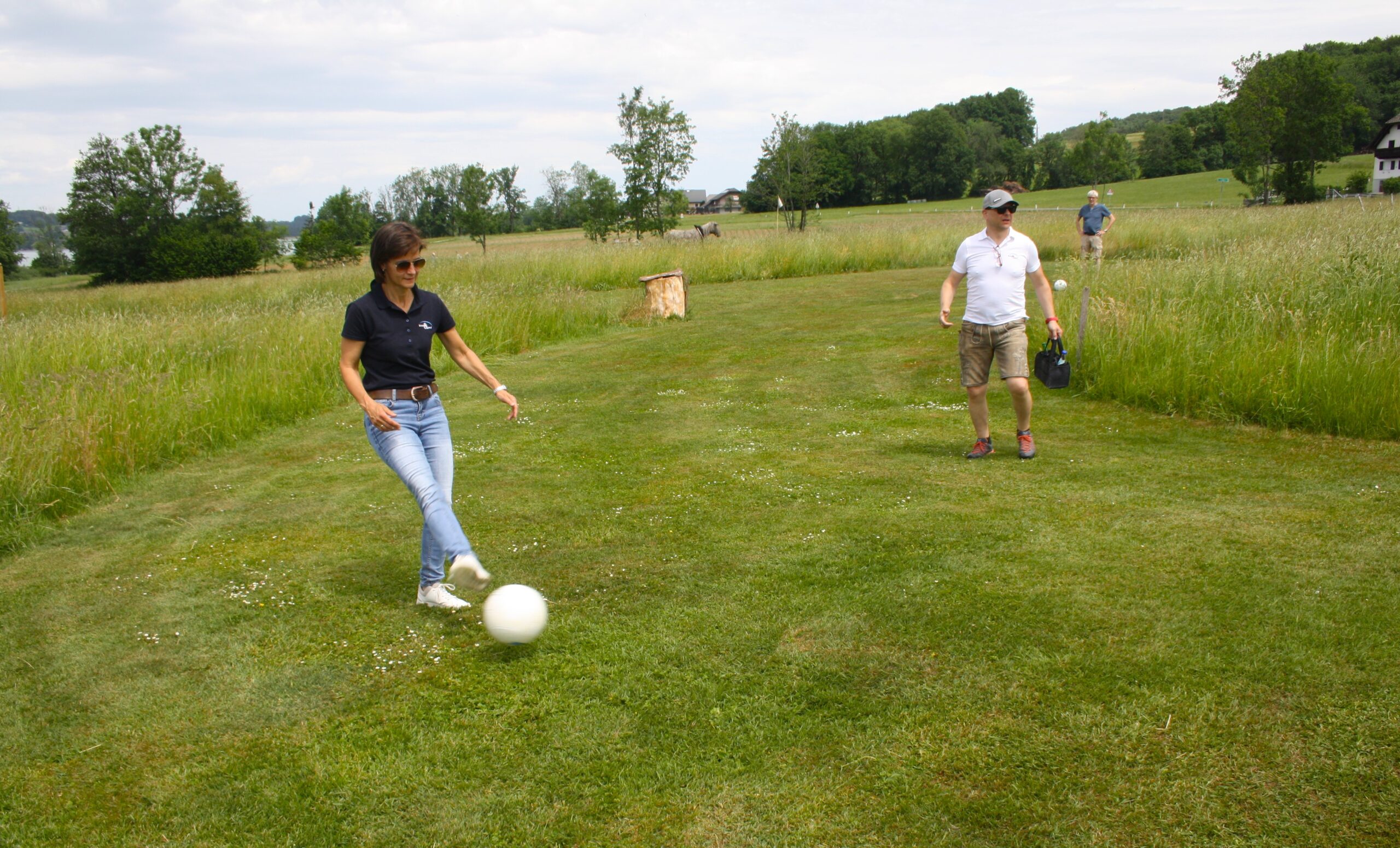 Eine Frau, die auf einer Wiese einen wei&szlig;en Ball kickt, w&auml;hrend zwei M&auml;nner zuschauen; im Hintergrund sind B&auml;ume und ein Haus zu sehen.