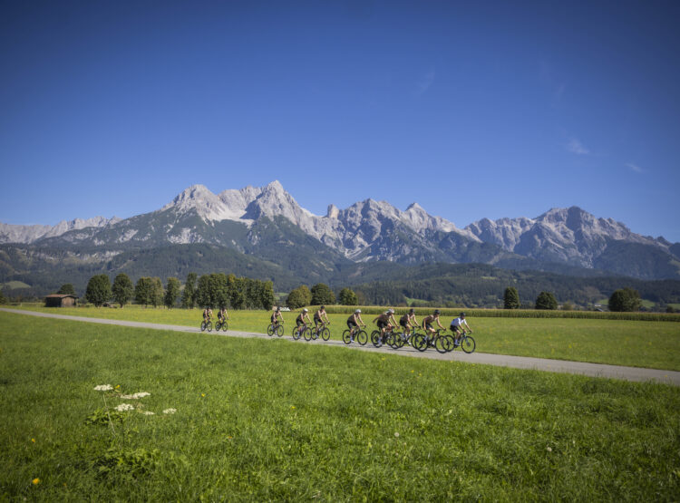 Eine Gruppe von Radfahrern fährt auf einem Weg durch eine grüne Wiese, im Hintergrund sind Berge zu sehen, und über ihnen erstreckt sich ein strahlend blauer Himmel.