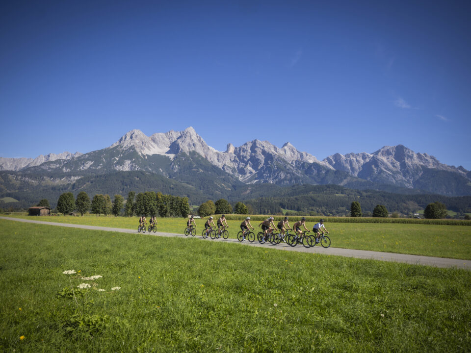 Eine Gruppe von Radfahrern fährt auf einem Weg durch eine grüne Wiese, im Hintergrund sind Berge zu sehen, und über ihnen erstreckt sich ein strahlend blauer Himmel.