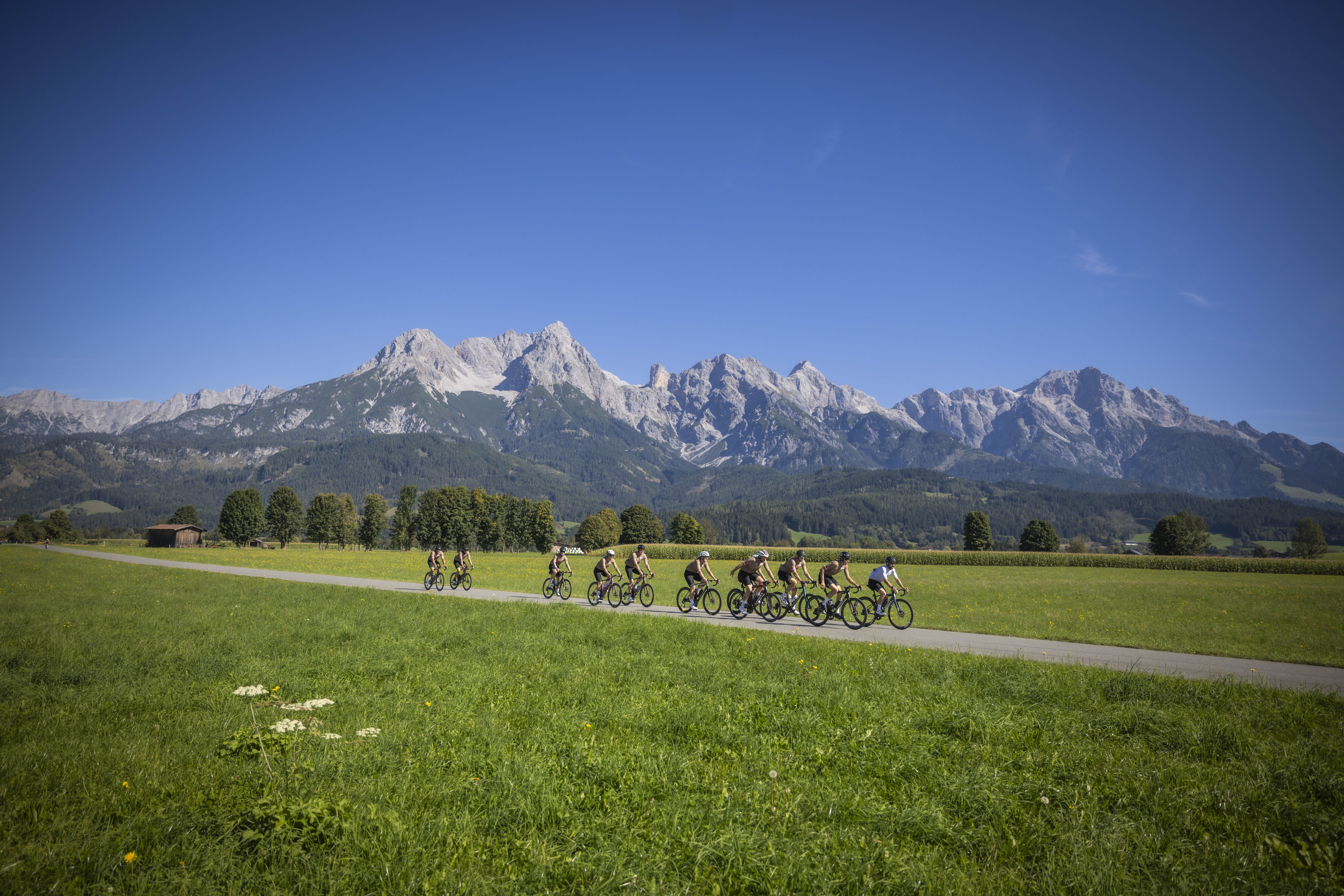 Eine Gruppe von Radfahrern fährt auf einem Weg durch grüne Felder mit Bergen im Hintergrund unter einem klaren blauen Himmel.
