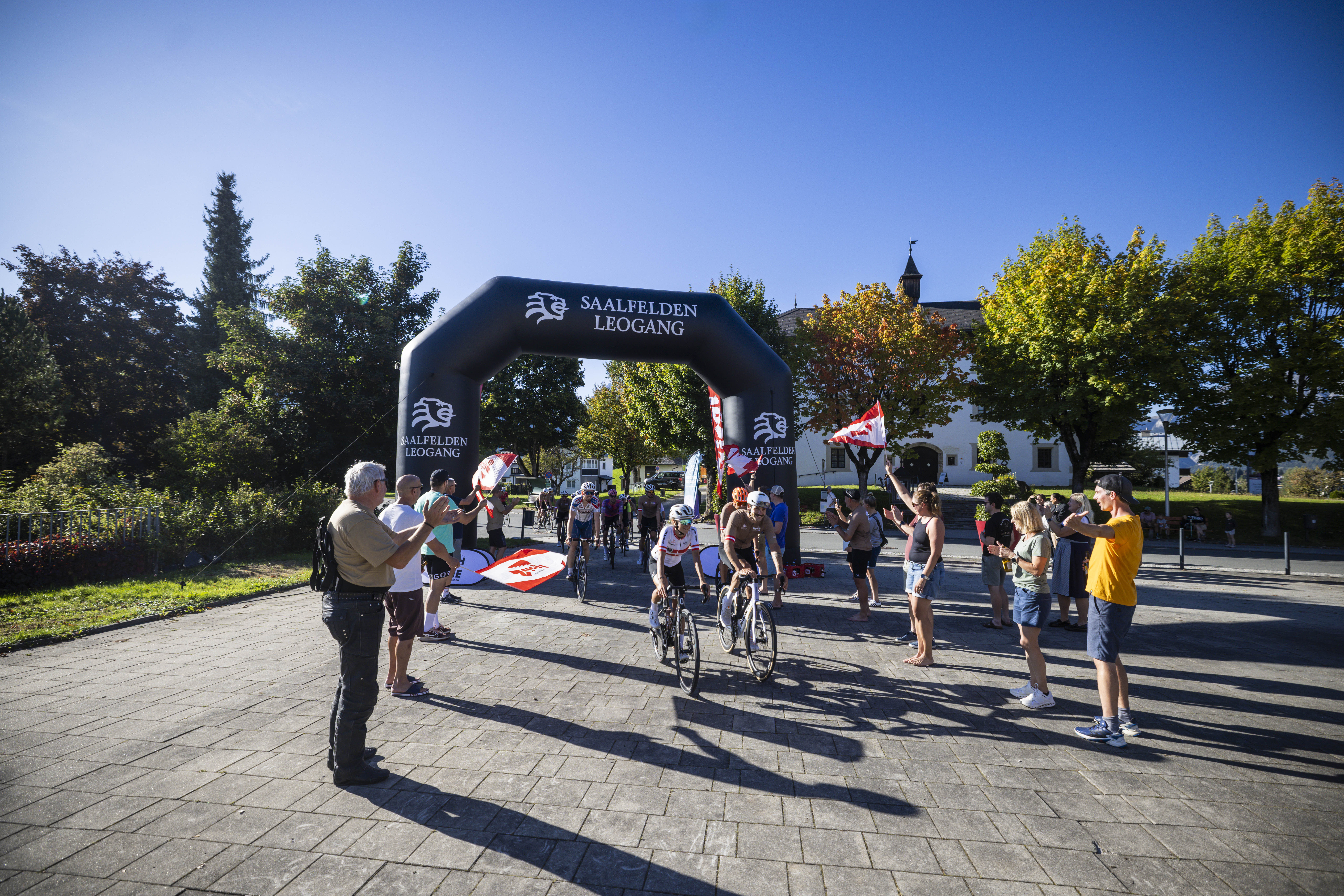 An einem sonnigen Tag beenden Radfahrer ein Rennen unter einem Torbogen, angefeuert von Menschen mit Fahnen und Kameras.