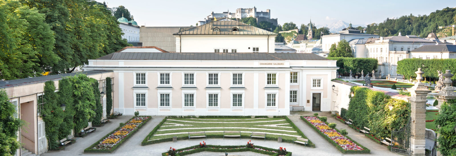 Ein formaler Garten mit bunten Blumenbeeten, einem Springbrunnen in der Mitte und einem weißen Gebäude im Hintergrund.