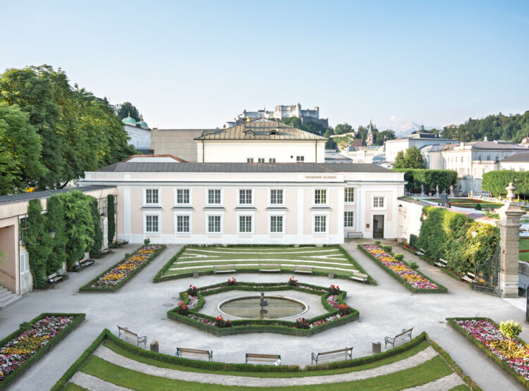 Ein formaler Garten mit bunten Blumenbeeten, einem Springbrunnen in der Mitte und einem weißen Gebäude im Hintergrund.
