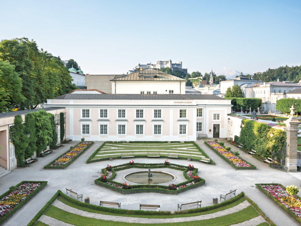 Ein formaler Garten mit bunten Blumenbeeten, einem Springbrunnen in der Mitte und einem weißen Gebäude im Hintergrund.