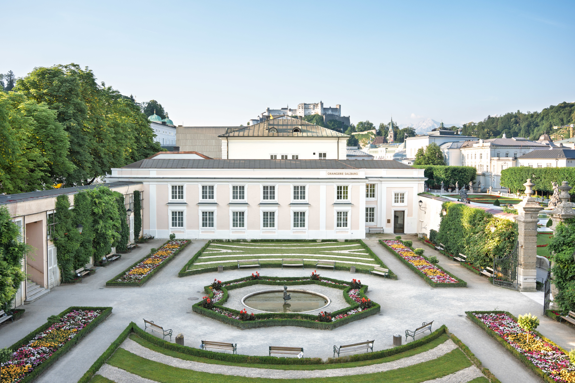 Ein formaler Garten mit bunten Blumenbeeten, einem Springbrunnen in der Mitte und einem weißen Gebäude im Hintergrund.
