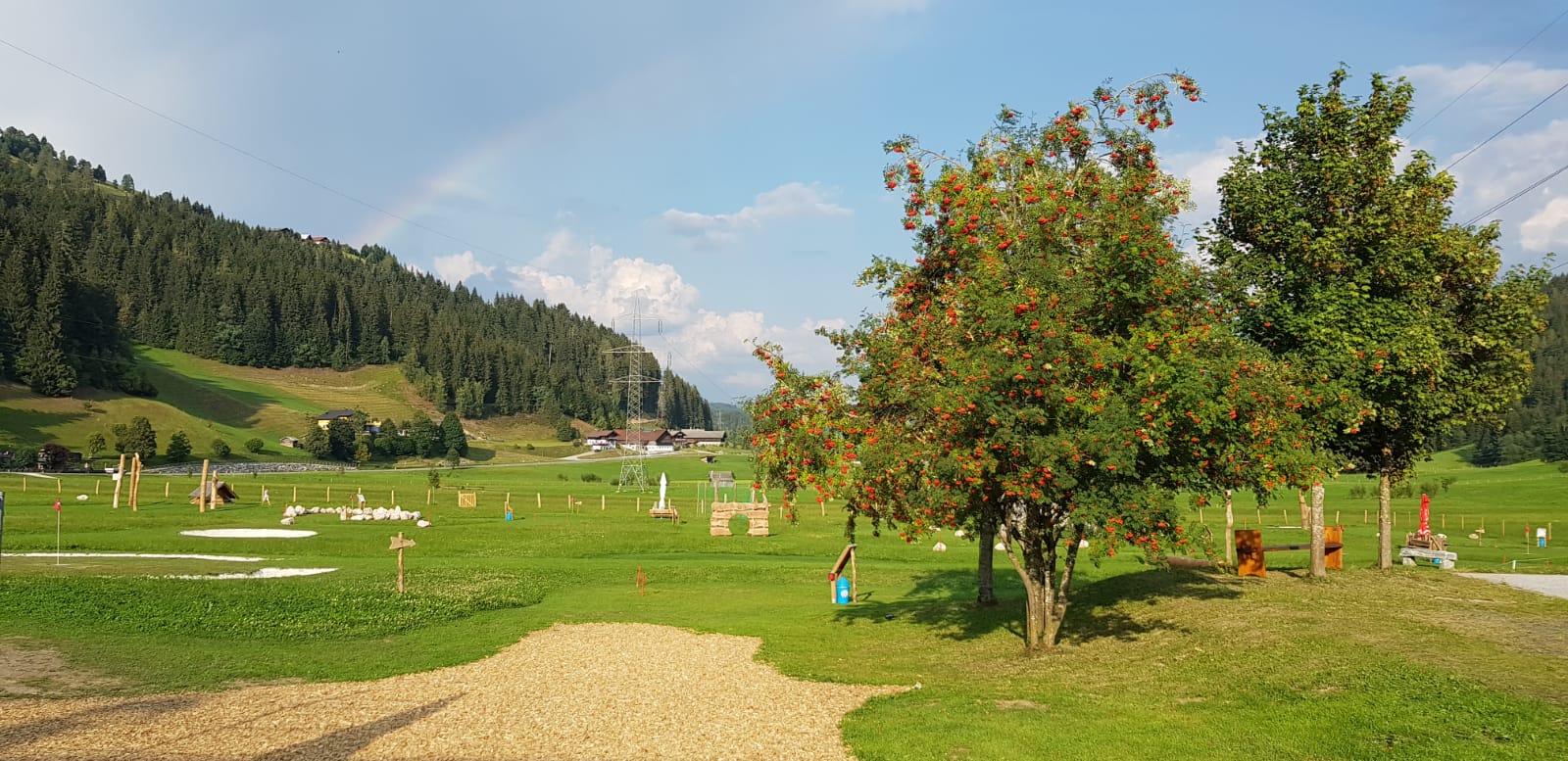 Eine gr&uuml;ne Wiese mit B&auml;umen, einigen vereinzelten Geb&auml;uden und einem schwachen Regenbogen am Himmel in der N&auml;he bewaldeter H&uuml;gel.