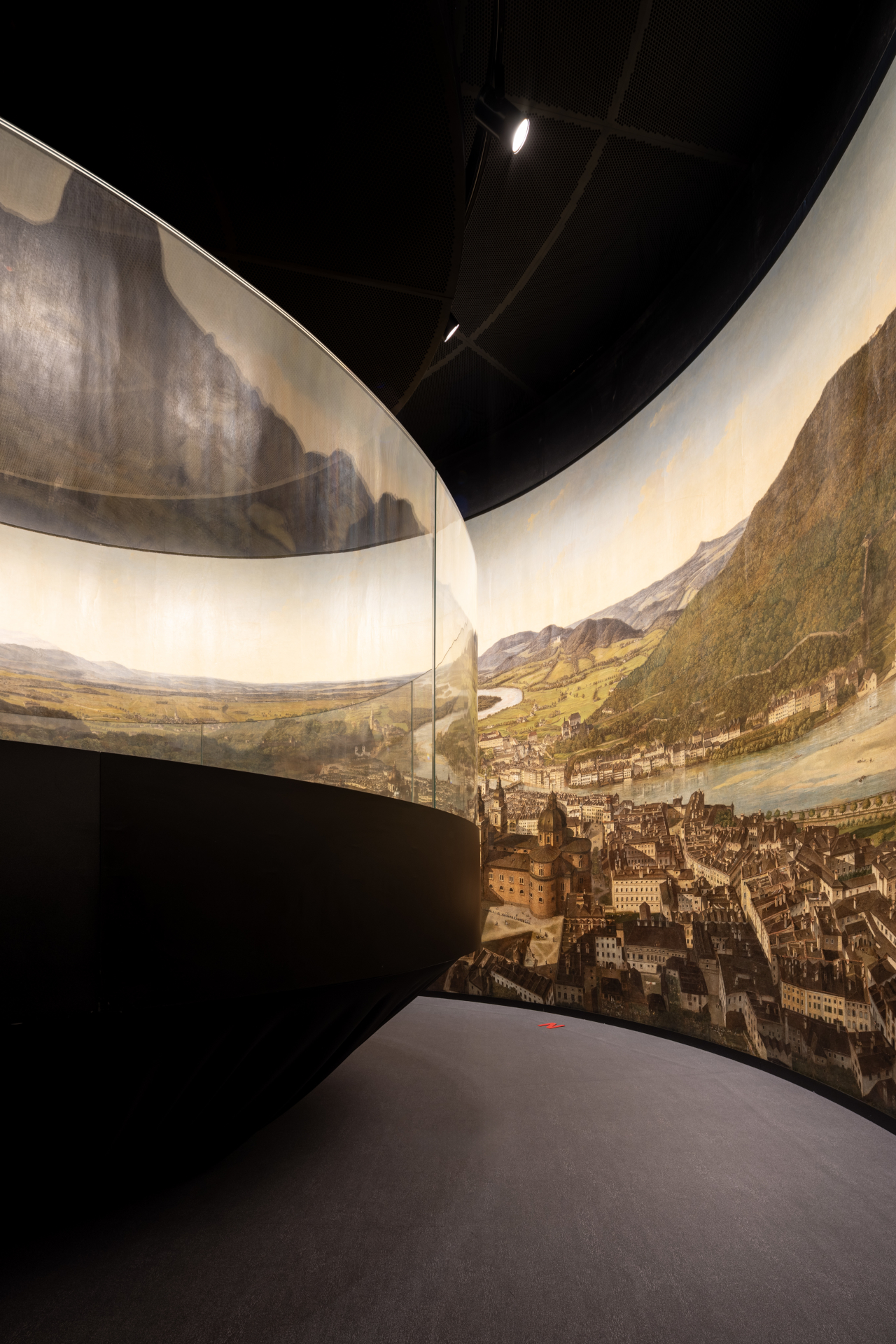 Ein großes Panoramagemälde, das eine Stadt und eine Landschaft zeigt und an geschwungenen Wänden in einer geräumigen, modernen Galerie ausgestellt ist.