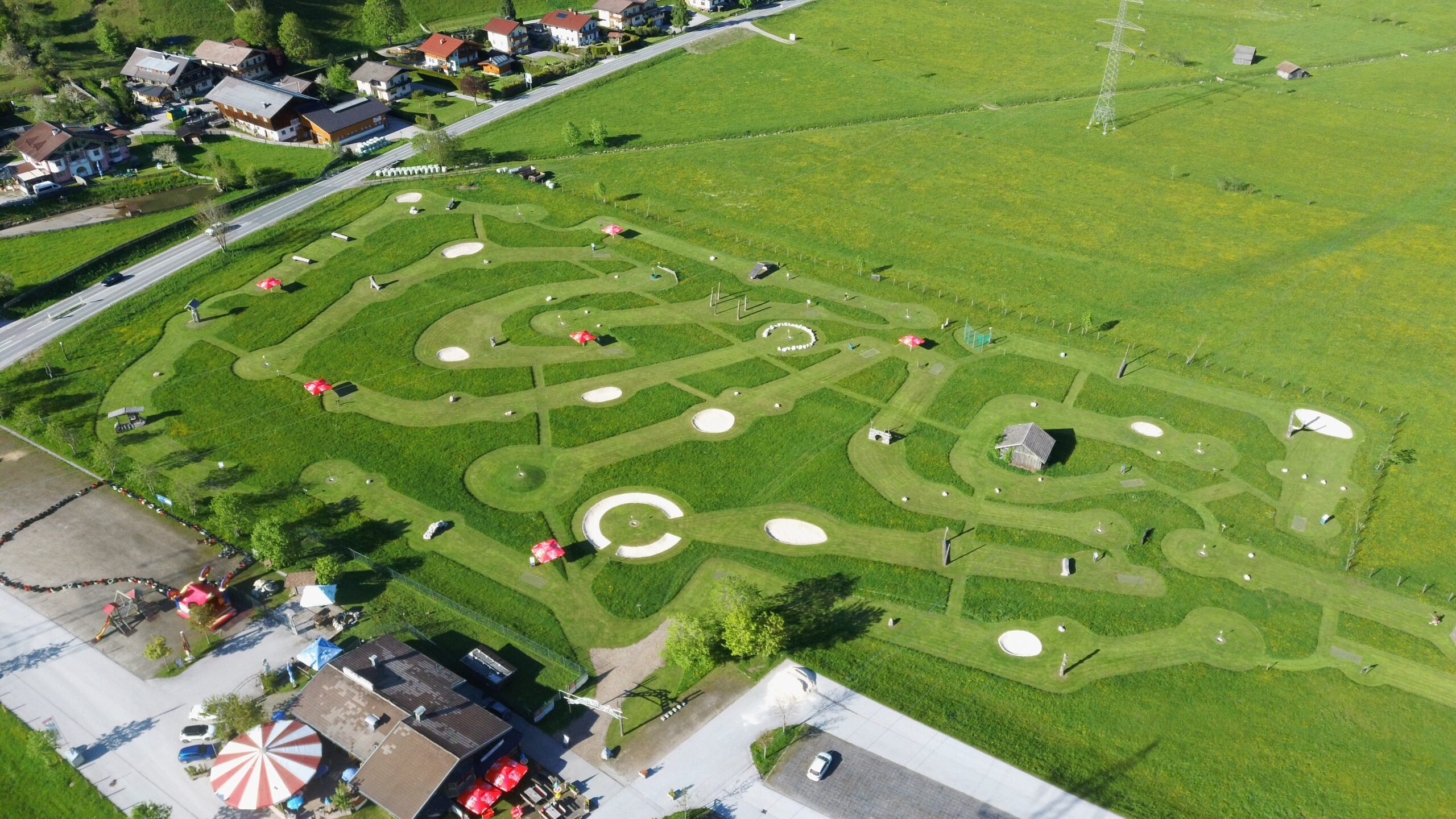Luftaufnahme eines Minigolfplatzes mit gewundenen Wegen, gr&uuml;nem Gras und vereinzelten roten Sonnenschirmen.