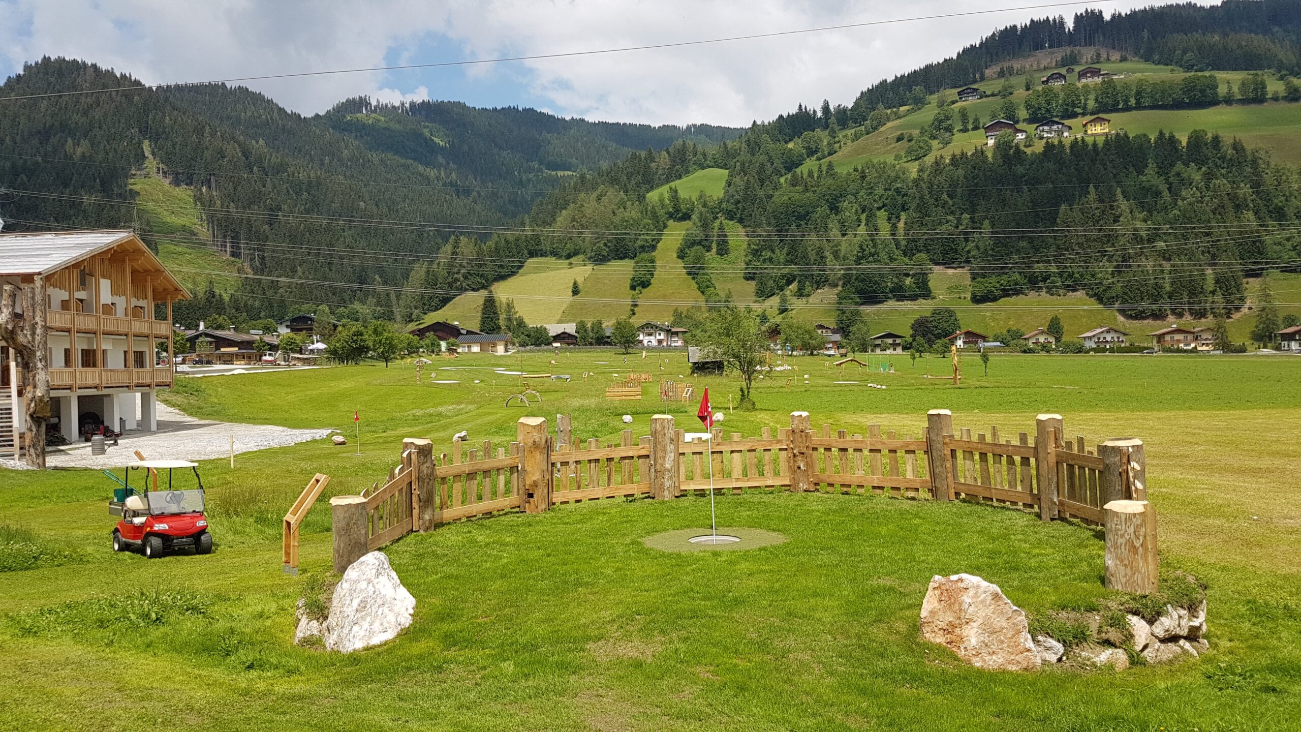 Ein runder Holzzaun auf einer Wiese mit H&uuml;geln, H&auml;usern und einem roten Golfwagen in der N&auml;he.