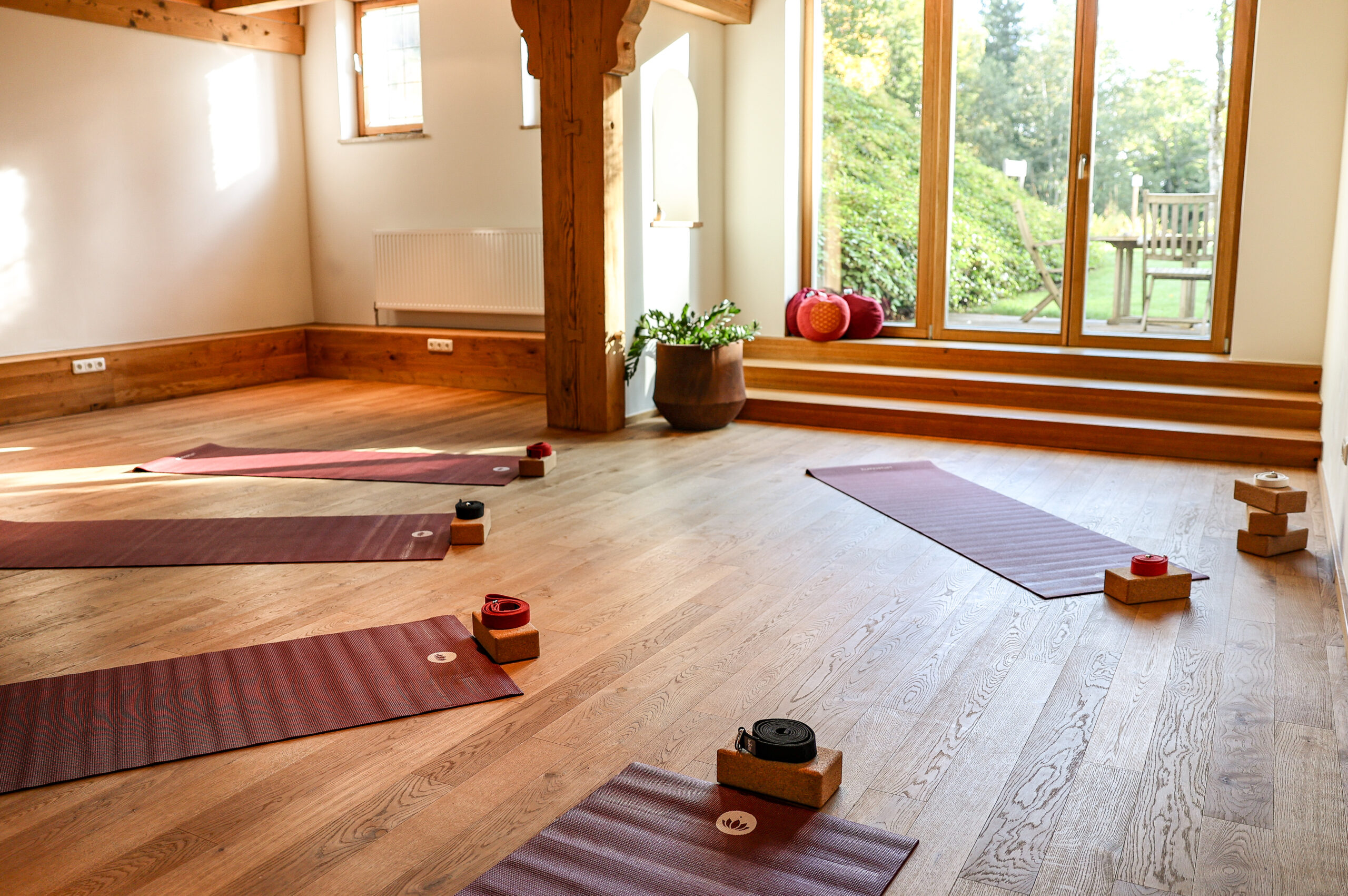 Ein helles Yogastudio mit Matten, Bl&ouml;cken und Gurten, die auf einem Holzboden in der N&auml;he gro&szlig;er Fenster und der gr&uuml;nen Umgebung drau&szlig;en ausgelegt sind.