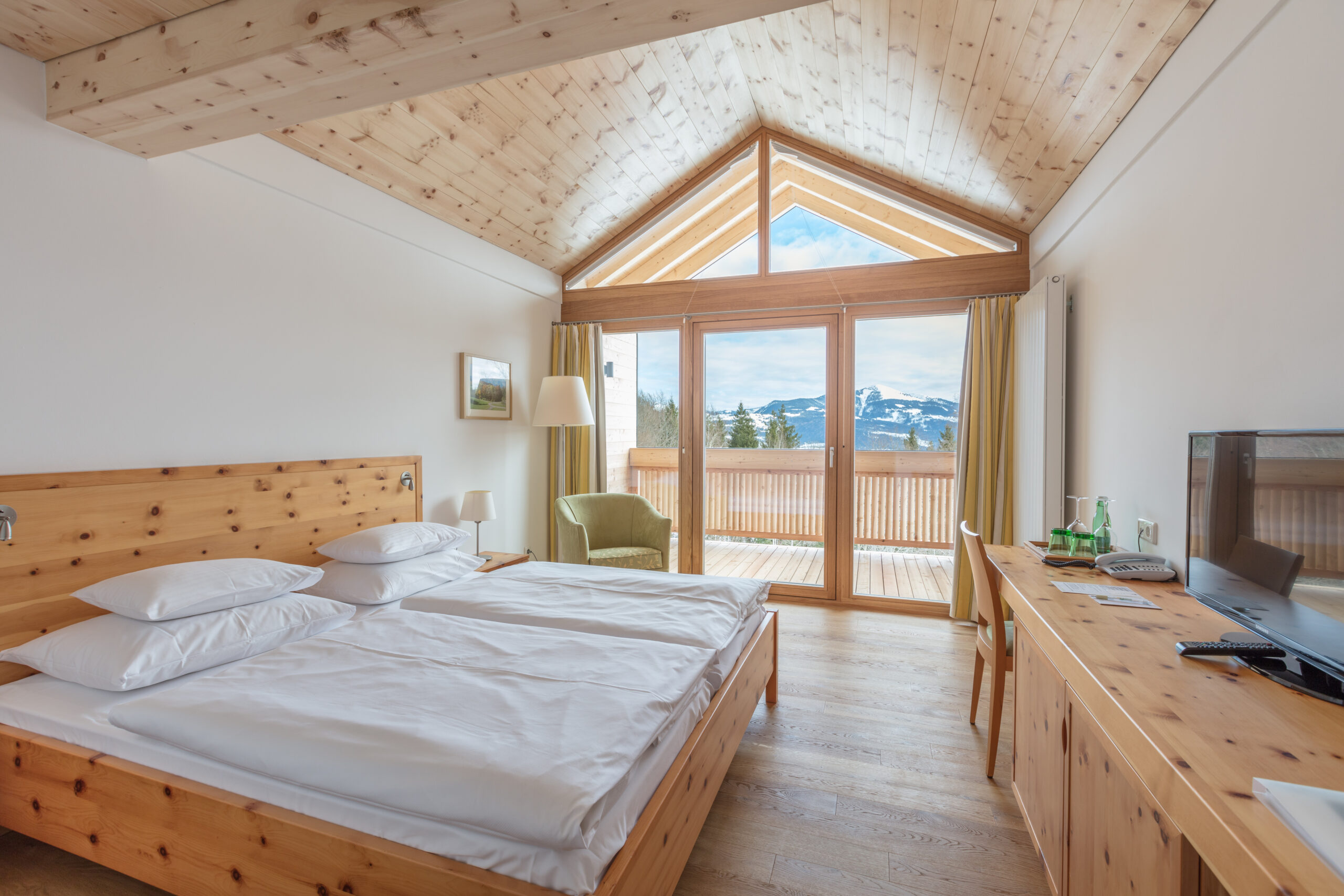 Ein gem&uuml;tliches Schlafzimmer aus Holz mit gro&szlig;en Fenstern, Blick auf die Berge, einem Doppelbett und einem Schreibtisch mit Fernseher.