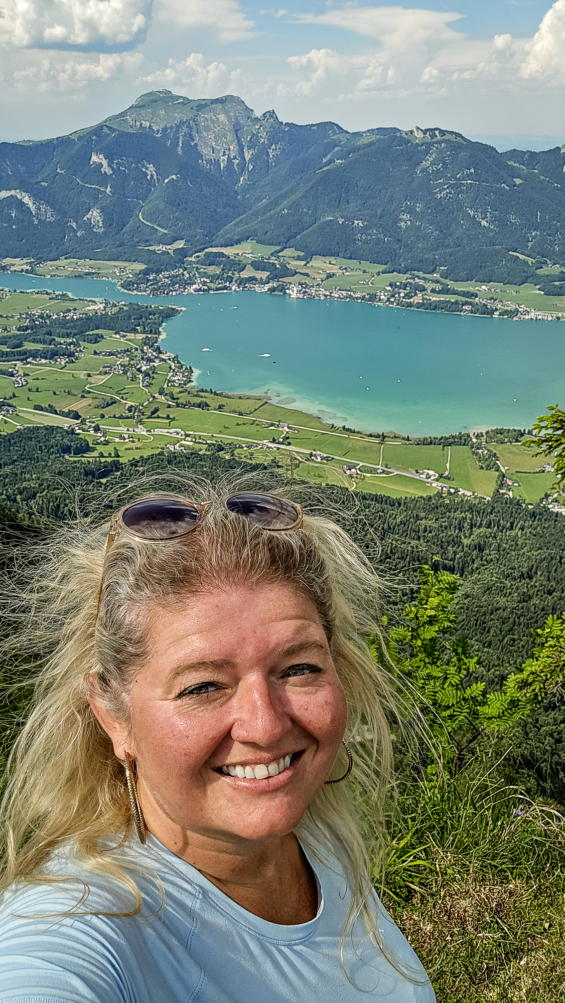 Eine l&auml;chelnde Frau macht ein Selfie vor dem Hintergrund der Bleckwandh&uuml;tte, eines Berges, eines Sees und einer gr&uuml;nen Tallandschaft.