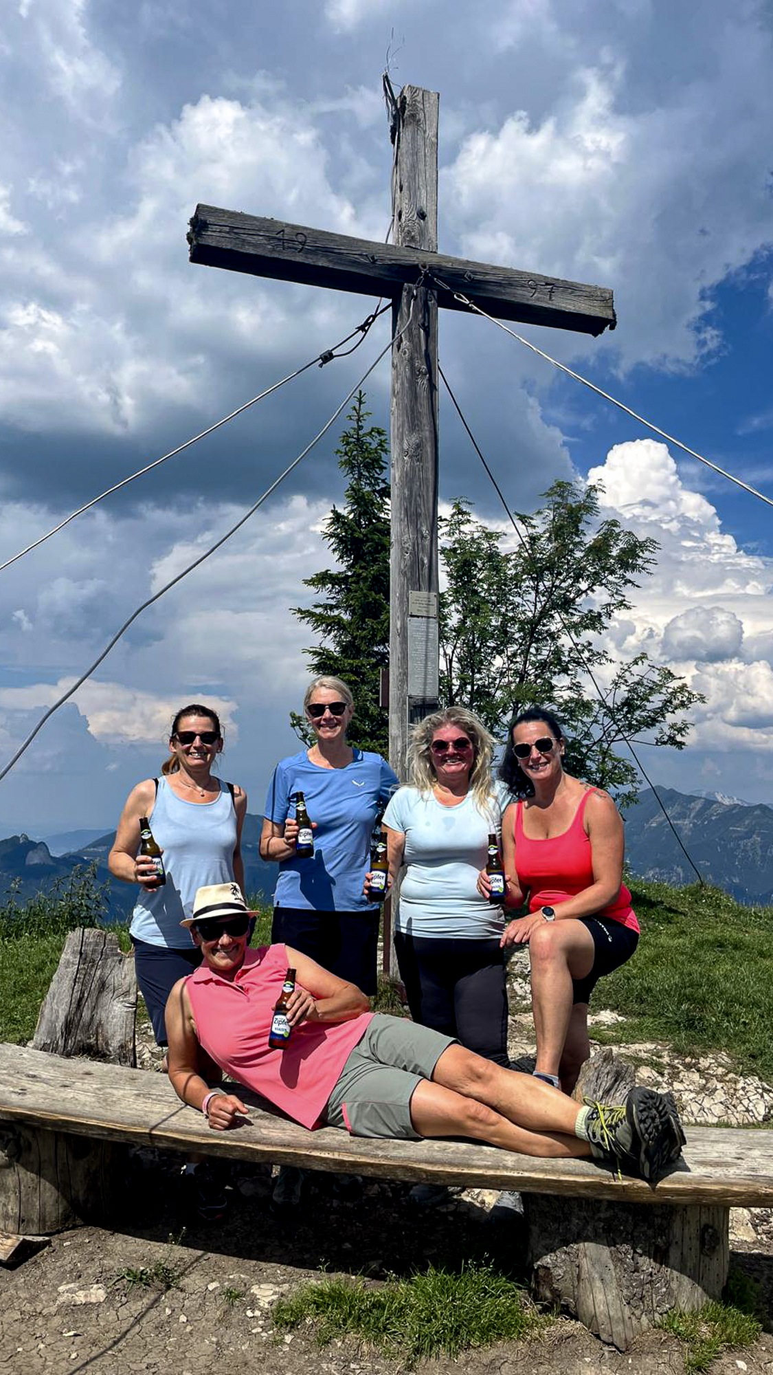 F&uuml;nf Personen mit Getr&auml;nken posieren unter teilweise bew&ouml;lktem Himmel neben einem gro&szlig;en Holzkreuz in der N&auml;he der Bleckwandh&uuml;tte.