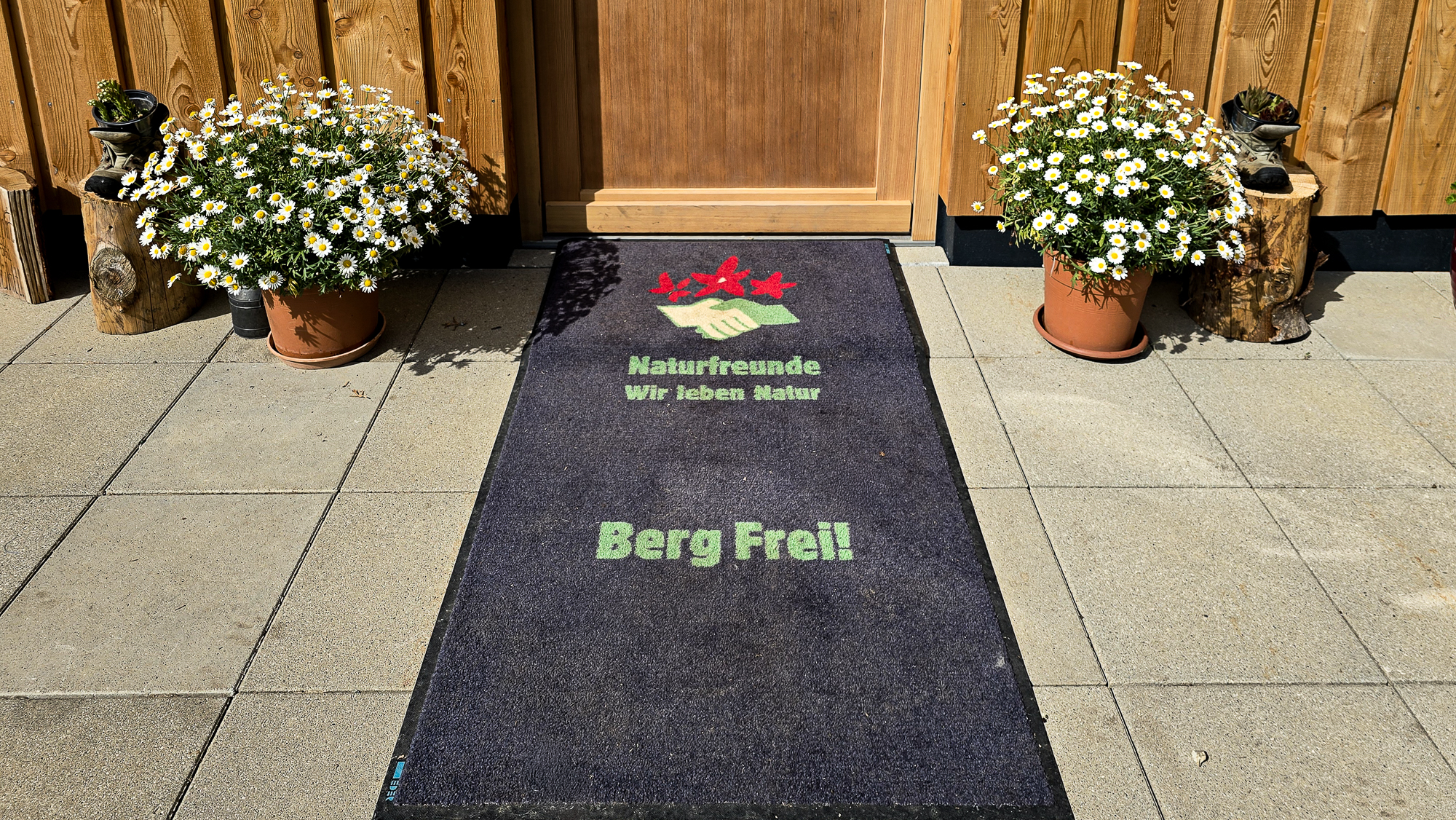 Fu&szlig;matte mit deutschem Text und Blument&ouml;pfen, die Sie vor einer Holzt&uuml;r in der Bleckwandh&uuml;tte willkommen hei&szlig;t.