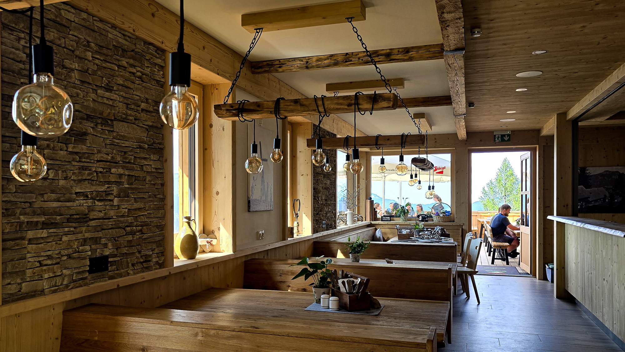 Gem&uuml;tliches Interieur der Bleckwandh&uuml;tte mit Holzm&ouml;beln, H&auml;ngeleuchten und einer Person, die am Fenster sitzt und den Blick ins Freie genie&szlig;t.
