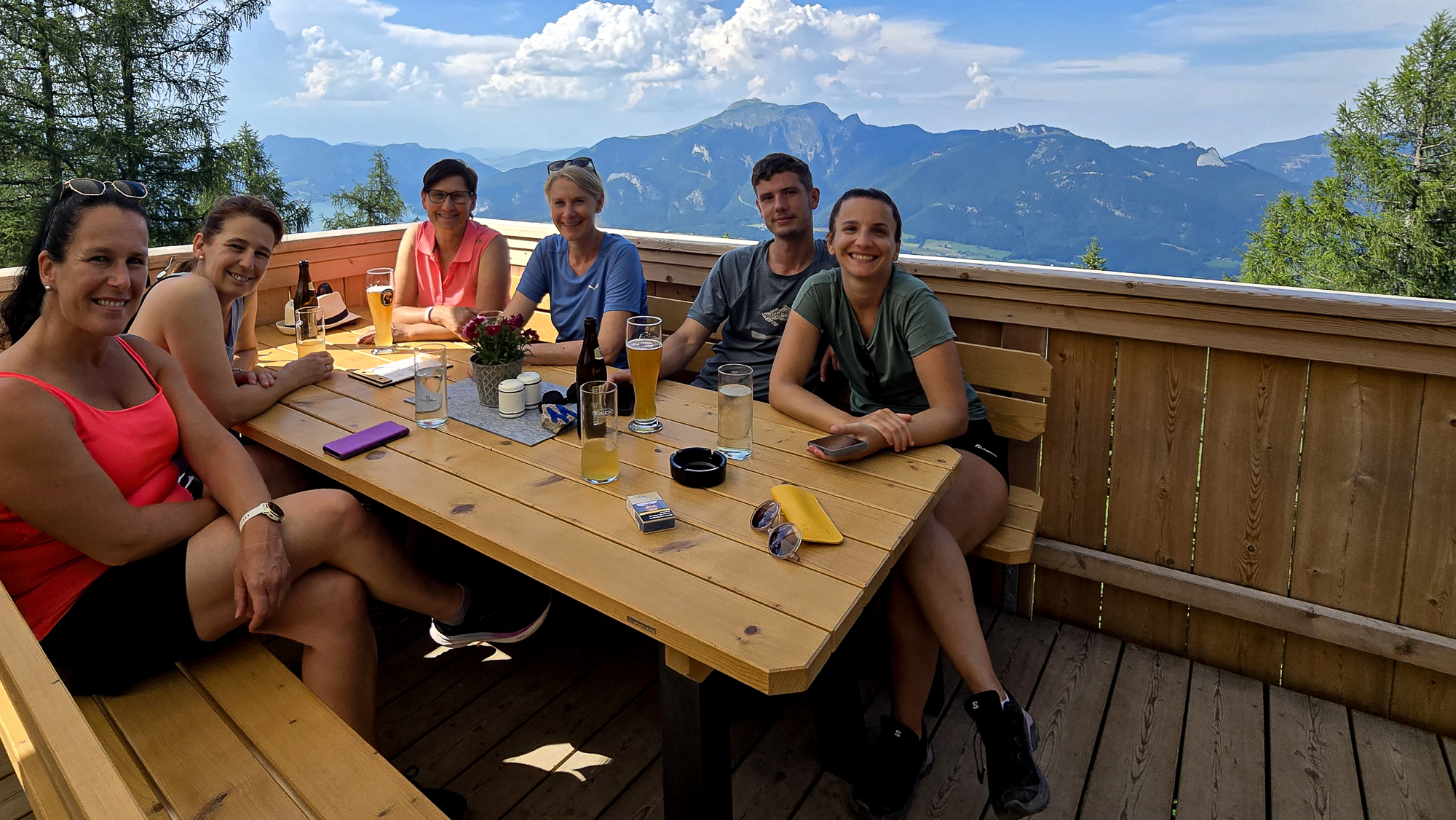 Sechs Personen sitzen an einem h&ouml;lzernen Picknicktisch in der Bleckwandh&uuml;tte und genie&szlig;en an einem sonnigen Tag die malerische Berglandschaft und ihre Getr&auml;nke.