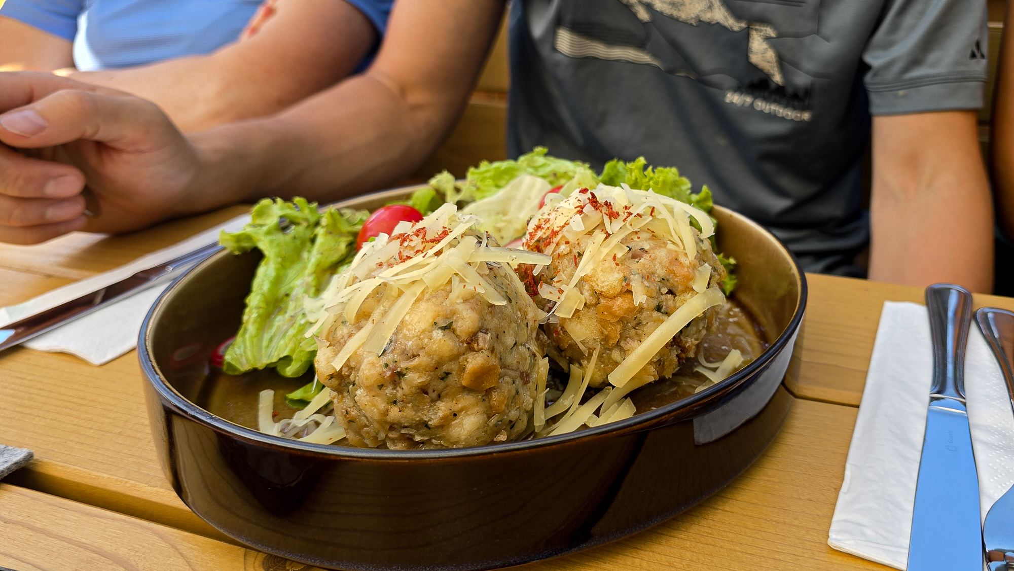 Zwei Personen sitzen an einem Tisch in der Bleckwandh&uuml;tte, vor sich Kn&ouml;del, geriebener K&auml;se, Salat und Tomaten.