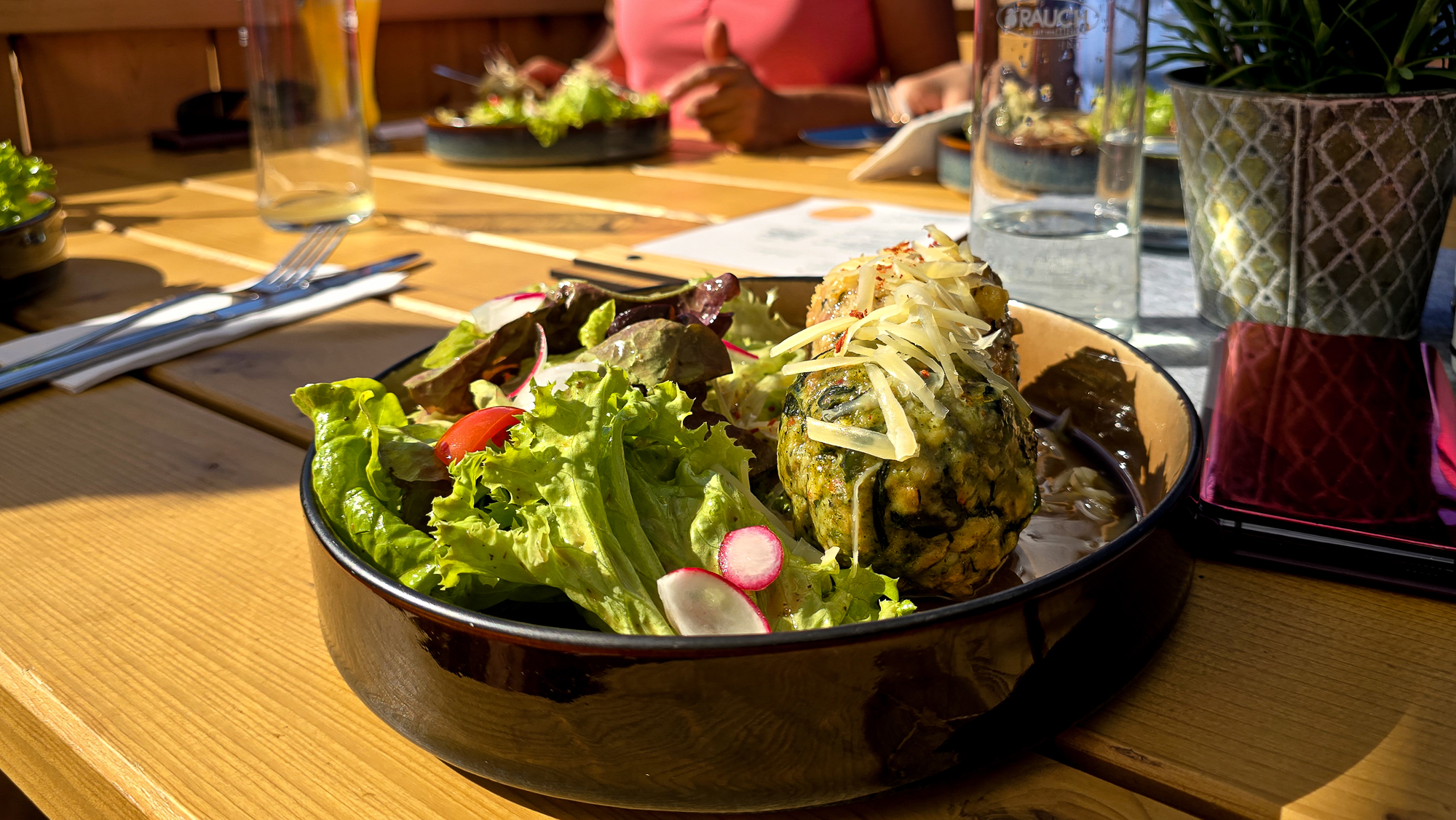 Eine Sch&uuml;ssel mit frischem Salat und Kn&ouml;deln in der Bleckwandh&uuml;tte, im Hintergrund sind G&auml;ste zu sehen, die im Freien essen.
