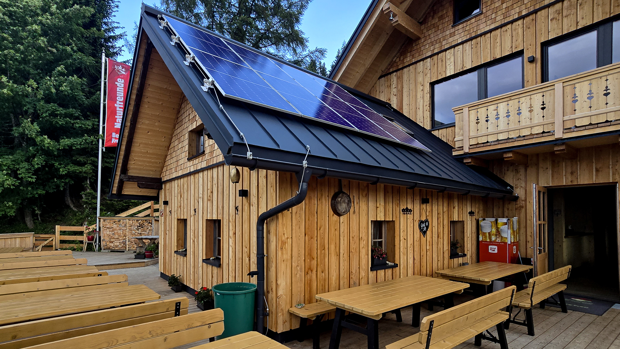 Bleckwandh&uuml;tte: Kleines Holzgeb&auml;ude mit Sonnenkollektoren, B&auml;nken im Freien und B&auml;umen im Hintergrund.