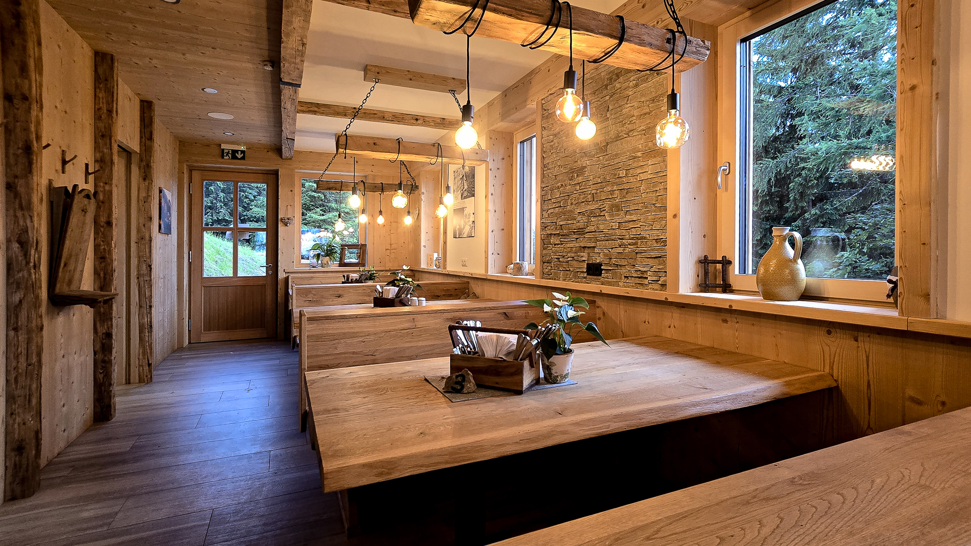 Gem&uuml;tliches Interieur der Bleckwandh&uuml;tte aus Holz mit Sitznischen, H&auml;ngeleuchten und gro&szlig;en Fenstern mit Blick auf den Wald.